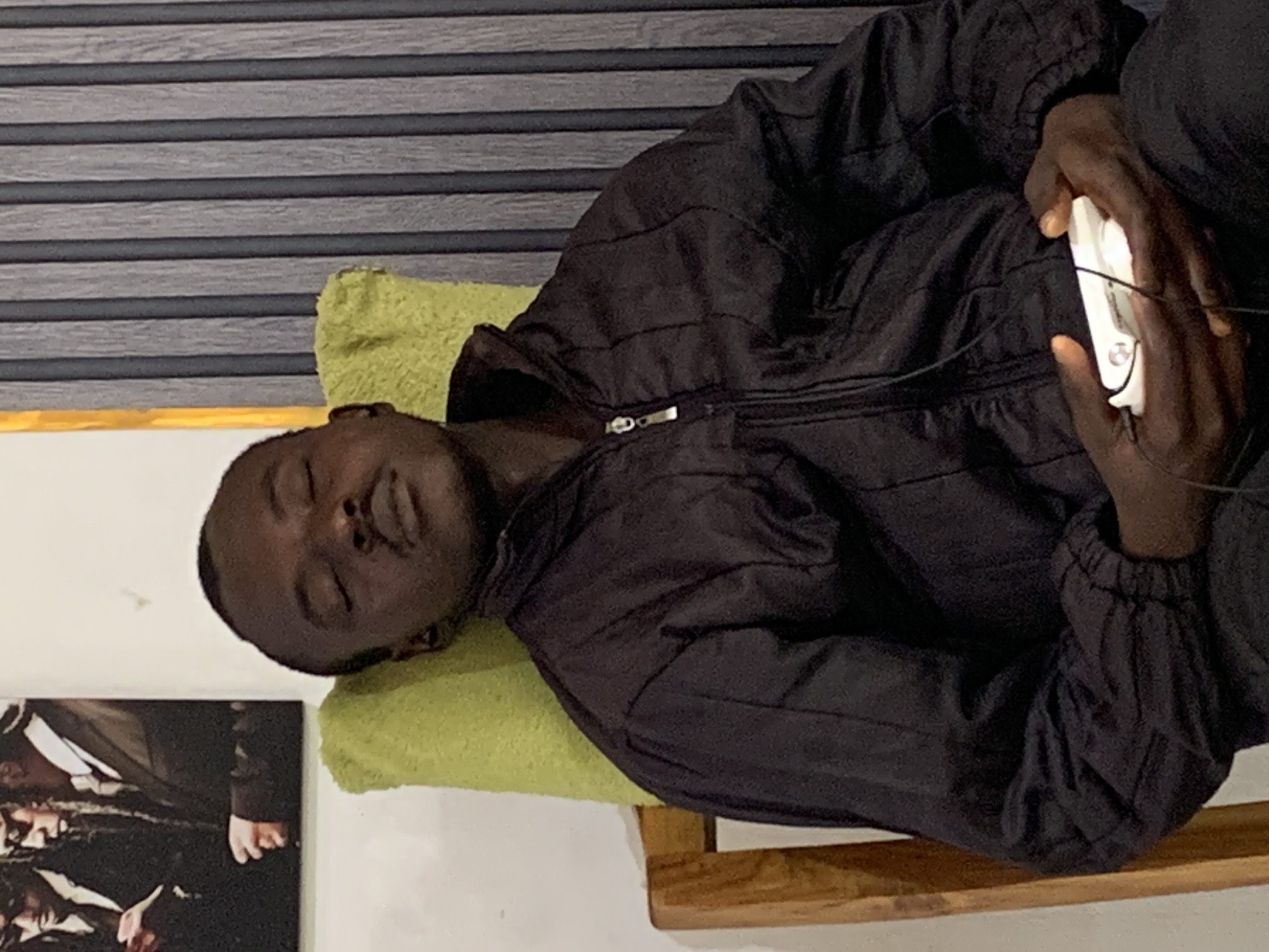 An African male is lying down on a bed. He is wearing a black puffy jacket. His head rests on a lime green pillow. He is holding a white electronic device in his hands, attached with a wire. A wooden frame supports the bed. In the background is a wall with a gray faux wood blind and a framed picture. This scene is in an indoor location in the city of Jos, Nigeria.