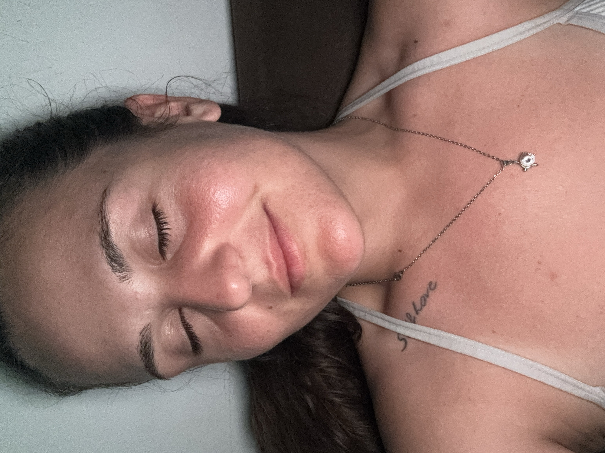 A close-up, shoulder-up shot captures a young woman with her eyes closed and a slight smile. Her head is tilted slightly to her left. She has medium-toned skin with visible texture and redness on her cheeks. Her dark hair is pulled back loosely, with a few strands falling around her face and neck. She is wearing a white halter top and a silver necklace with a small, sparkling pendant. A tattoo, partially visible on her left shoulder, reads "still love". The background is a plain, neutral wall, suggesting an indoor setting. There is no information in the image that indicates a specific location in Plovdiv, Bulgaria.