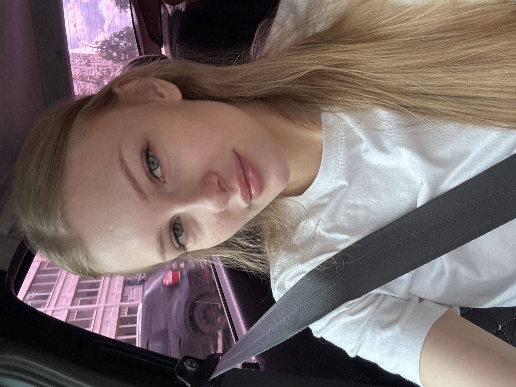 A young woman with blonde hair and blue eyes, wearing a white t-shirt and a seatbelt, is pictured in the passenger seat of a car. She is looking at the camera, and her lips are slightly parted. The background shows a building with many windows, possibly an apartment building, and trees. The overall lighting suggests it is daytime, and the pinkish hue in the background might be due to tinted windows or the time of day. The setting is likely a street in the city of Perm, Russia.