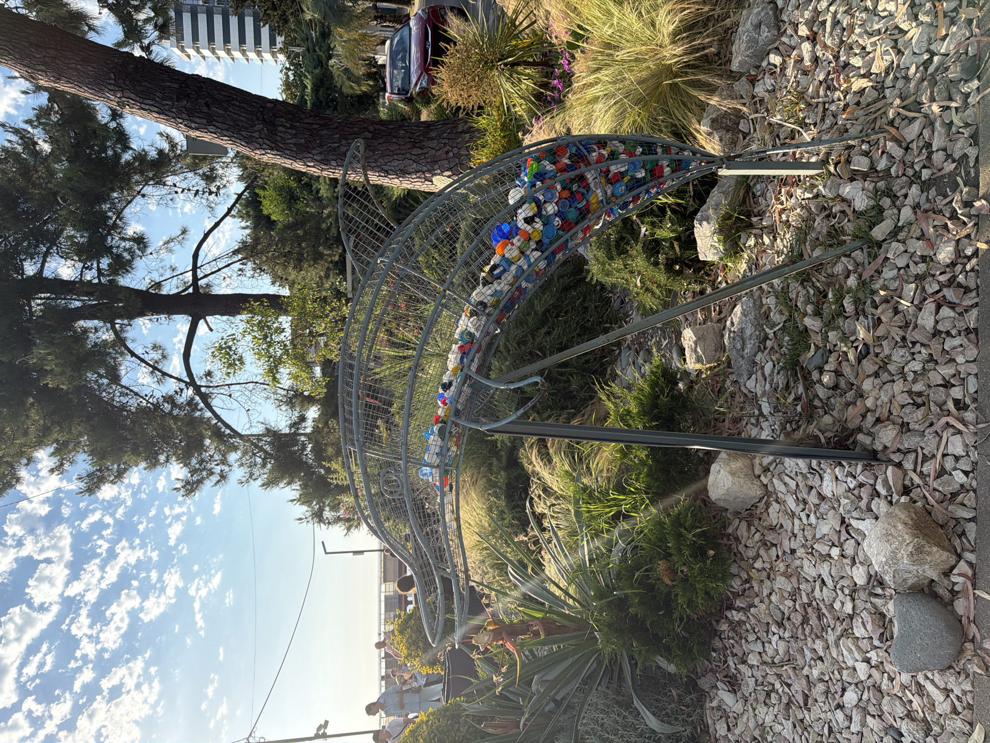This is a wide-angle, eye-level photograph taken outdoors in what appears to be a public park or landscaped area in Sochi, Russia, with the Black Sea visible in the background. The sky is bright blue with scattered white clouds. In the foreground, a wire sculpture in the shape of a dolphin is filled with colorful plastic bottle caps. The sculpture is supported by two metal legs and stands amidst dense green foliage and gravel. Two people are visible in the mid-ground, standing near a railing on a promenade. A red car is parked on a street in the background, beyond a large pine tree. The overall lighting suggests it is late afternoon or early evening.