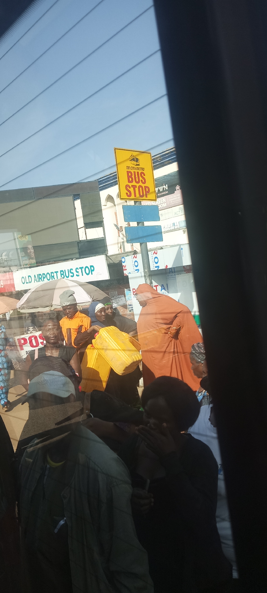 The media file captures a busy daytime street scene in Jos, Nigeria, viewed from inside a vehicle, likely through a windshield with reflections and defroster lines. The prominent feature is a yellow sign indicating "TIN CITY METRO BUS STOP," further confirmed by a white sign below it reading "OLD AIRPORT BUS STOP."
Several individuals are gathered on what appears to be a sidewalk or street. In the foreground, a woman in a black top looks towards the camera. Beside her, a man in an orange shirt and grey beanie stands near a woman holding a large yellow plastic jerrycan. Another person in a distinctive burnt-orange hooded garment is visible further to the right. An umbrella provides shade in the mid-ground. The presence of the jerrycan suggests daily activities such as fetching water or other liquids.
The background features various commercial signs and billboards, some partially obscured or blurred. Visible text includes "MULTIMEDIA," "GREEN DOM," "ACT," "TAI," and "JAL." There's also a partial "POS" sign. The clear sky and bright lighting indicate it is daytime. The overall impression is a bustling urban environment characteristic of a public transport hub.