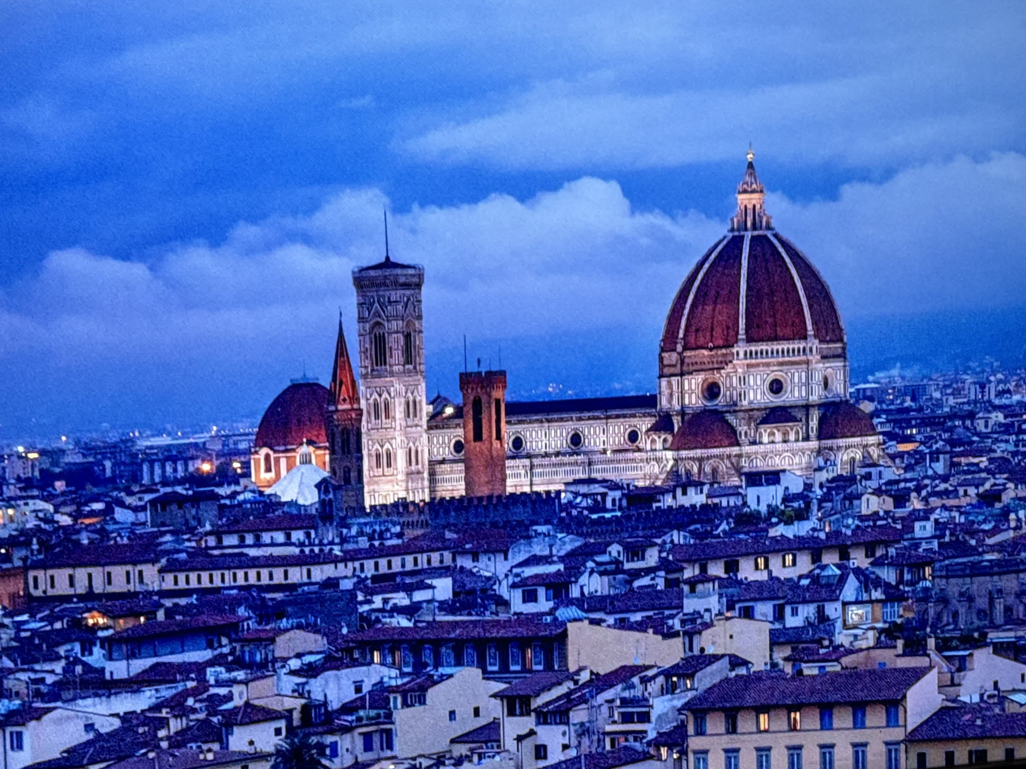 This is a nighttime aerial view of Florence, Italy, featuring the Duomo (Cathedral of Santa Maria del Fiore) prominently in the center. The iconic red-domed cathedral is illuminated, with its bell tower (Giotto's Campanile) to its left. The foreground shows a dense collection of terracotta-tiled rooftops of the city's buildings, indicating an urban environment. The sky is a deep blue, with some scattered clouds, suggesting dusk or early night. There are no people, animals, or specific actions visible in the image. The scene is a static cityscape. There is no depiction of Apostadero, Venezuela in this image.