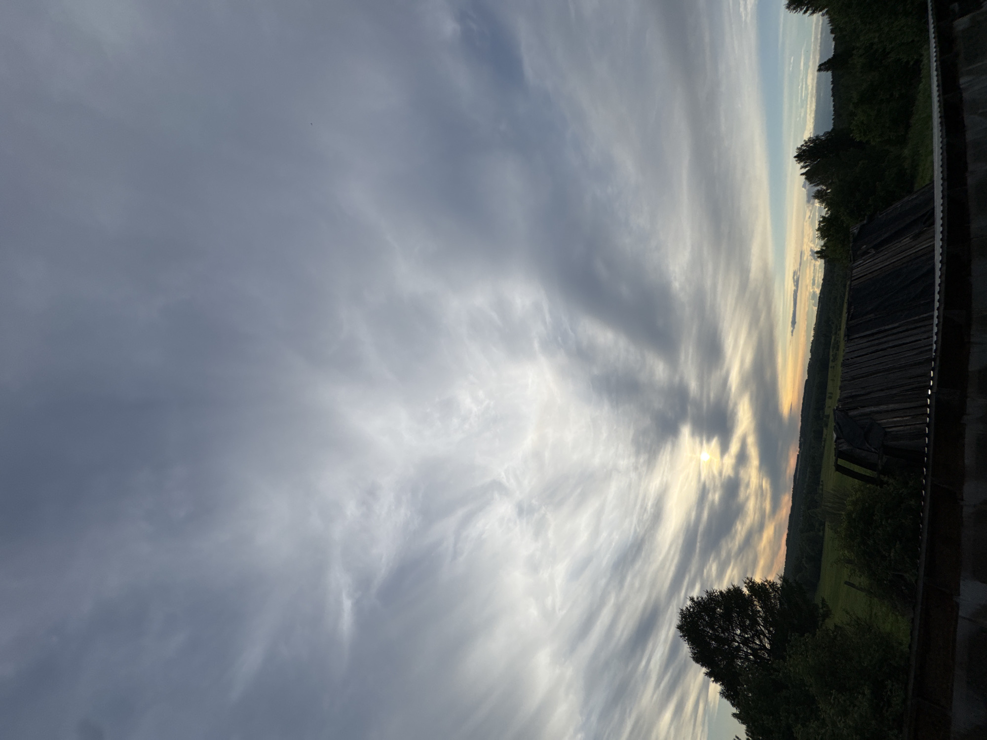 The image captures a landscape scene in Nizhnyaya Gar', Russia, with a prominent sky filled with dynamic, elongated clouds stretching across the frame. The clouds appear to radiate from a central point where the sun is partially visible, casting a soft, warm glow. The horizon line is marked by a dense forest area. To the right, there is a wooden structure, possibly a building or fence, partially visible and providing a sense of depth to the scene. The foreground is lined with greenery, including trees and shrubs. The overall atmosphere suggests a tranquil and possibly rural environment.