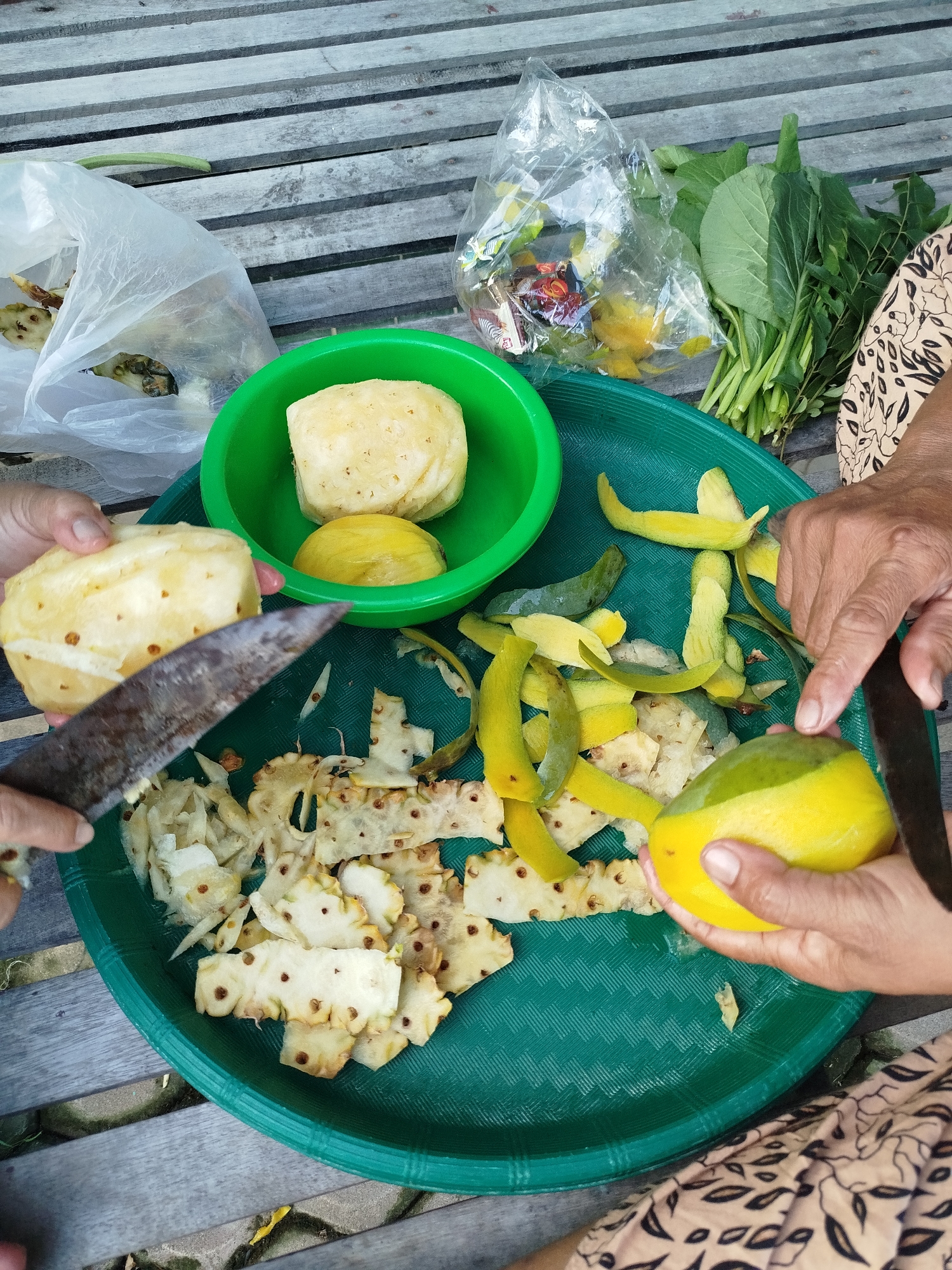 The image, likely taken in Tanjonghajimuda, Indonesia, shows a person peeling fruit. The setting appears to be an outdoor wooden bench, upon which a green plastic tray holds pineapple and mango. A portion of the pineapple has been peeled, and discarded rinds and peeled pieces are visible on the tray. A person's hands are holding a half-peeled mango and a knife. Another green bowl containing peeled fruit sits on the tray. There are bags and what appears to be leafy greens in the background. The scene depicts the act of preparing fruit for consumption.