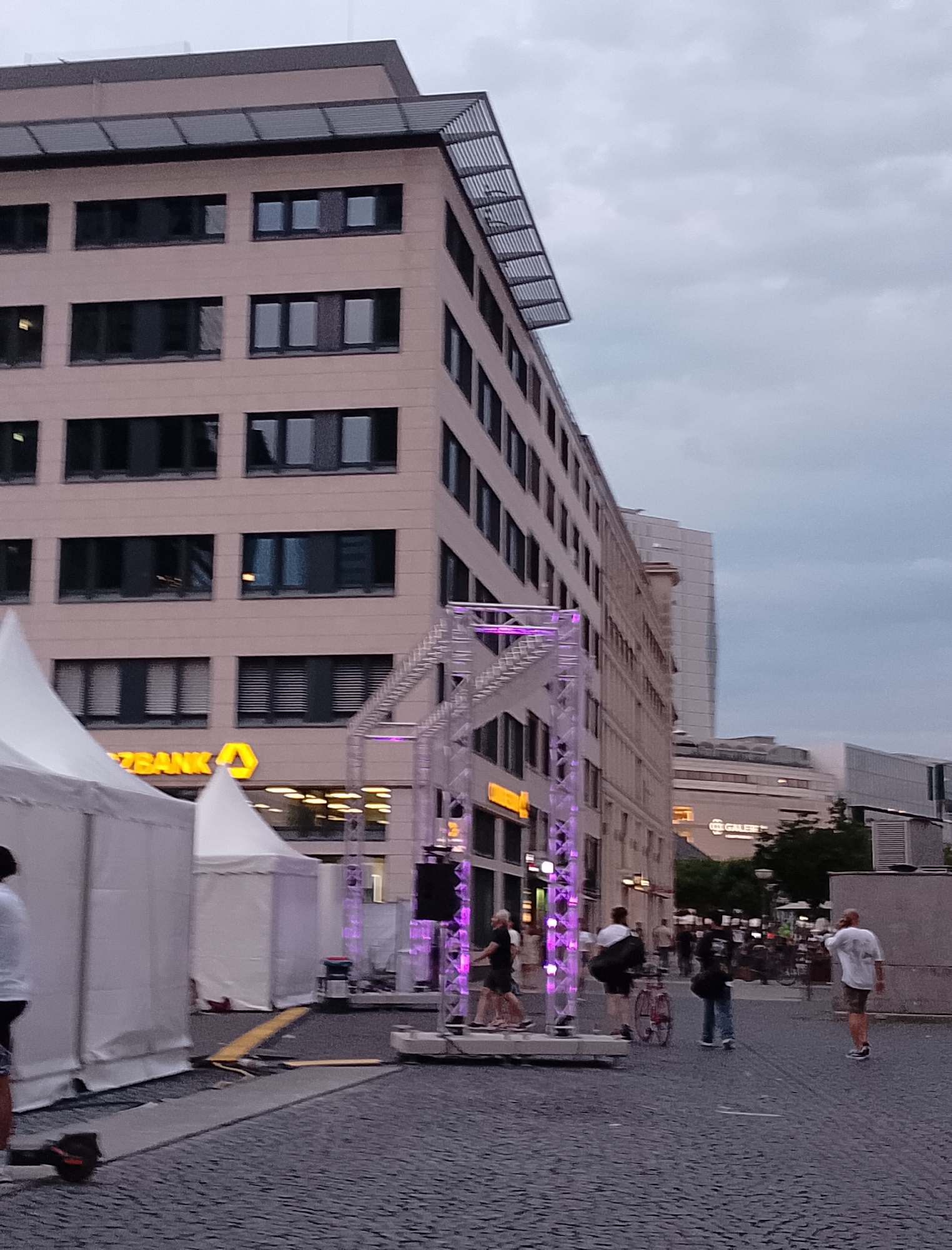 The image depicts a street scene in an urban environment, possibly during dusk or an overcast day. In the foreground, a dark cobblestone pavement is visible, with a section of what appears to be a temporary walkway or ramp. To the left, a large white tent is partially visible. Further into the scene, a light-colored, modern building with many windows dominates the background. The building has a dark, slatted roof structure. Adjacent to this building, a metallic truss structure, illuminated with purple lights, stands prominently.
Several people are present, walking in various directions. Some are seen from behind, while others are in profile. One person on the left is wearing a white shirt and dark shorts, and another further into the scene is wearing a white shirt and khaki shorts, holding a phone to their ear. A person in a black jacket and blue jeans is walking away from the camera, and a person with a red bicycle is also present. Tents, possibly for an event or market, are set up on the left side of the image. In the middle ground, another building with illuminated signage is visible, including the word "BANK" and a yellow logo. Other buildings and streetlights are present in the distance. The sky is cloudy and grey.
