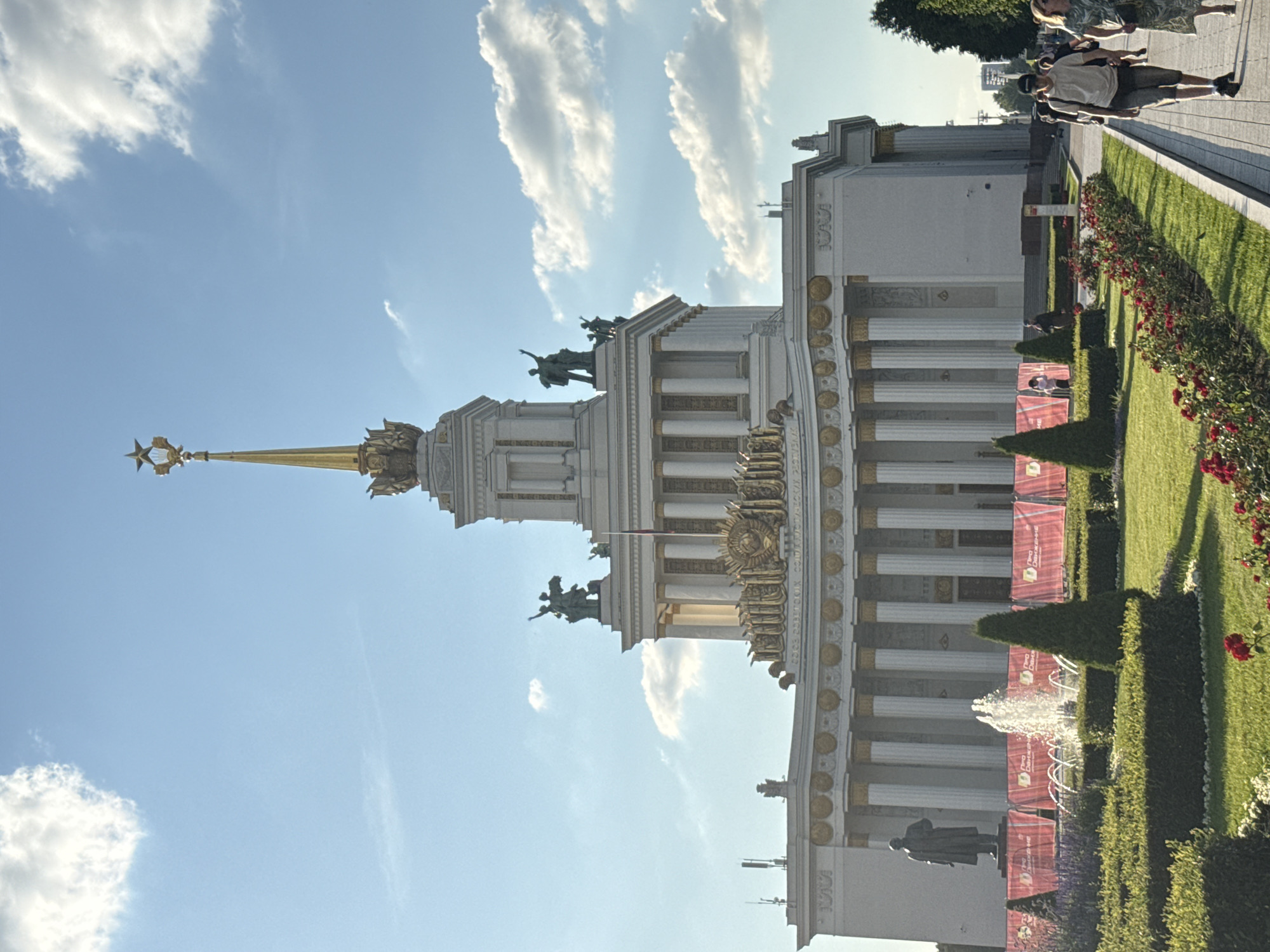 The image captures a wide-angle view of the main pavilion at VDNKh (Exhibition of Achievements of National Economy) in Moscow, Russia. The monumental building, with its distinctive Stalinist architecture, is a central focus, topped by a golden spire and star. Statues adorn the upper levels of the structure. In the foreground, a well-maintained garden with manicured hedges, green grass, and blooming red roses is visible. A fountain is active in the garden. To the left of the fountain, a dark statue of a man stands. Red banners with text are displayed behind the garden. A paved walkway runs along the right side of the image, where several individuals are walking. On the walkway, a man in a light-colored t-shirt and shorts, wearing a baseball cap, is walking towards the viewer. Next to him, a woman in a patterned dress is walking away from the viewer. The sky above is blue with scattered white clouds, suggesting a bright, possibly late afternoon, day. Trees line the right edge of the walkway and extend into the background. The overall scene conveys a sense of a public park or exhibition space with architectural grandeur.