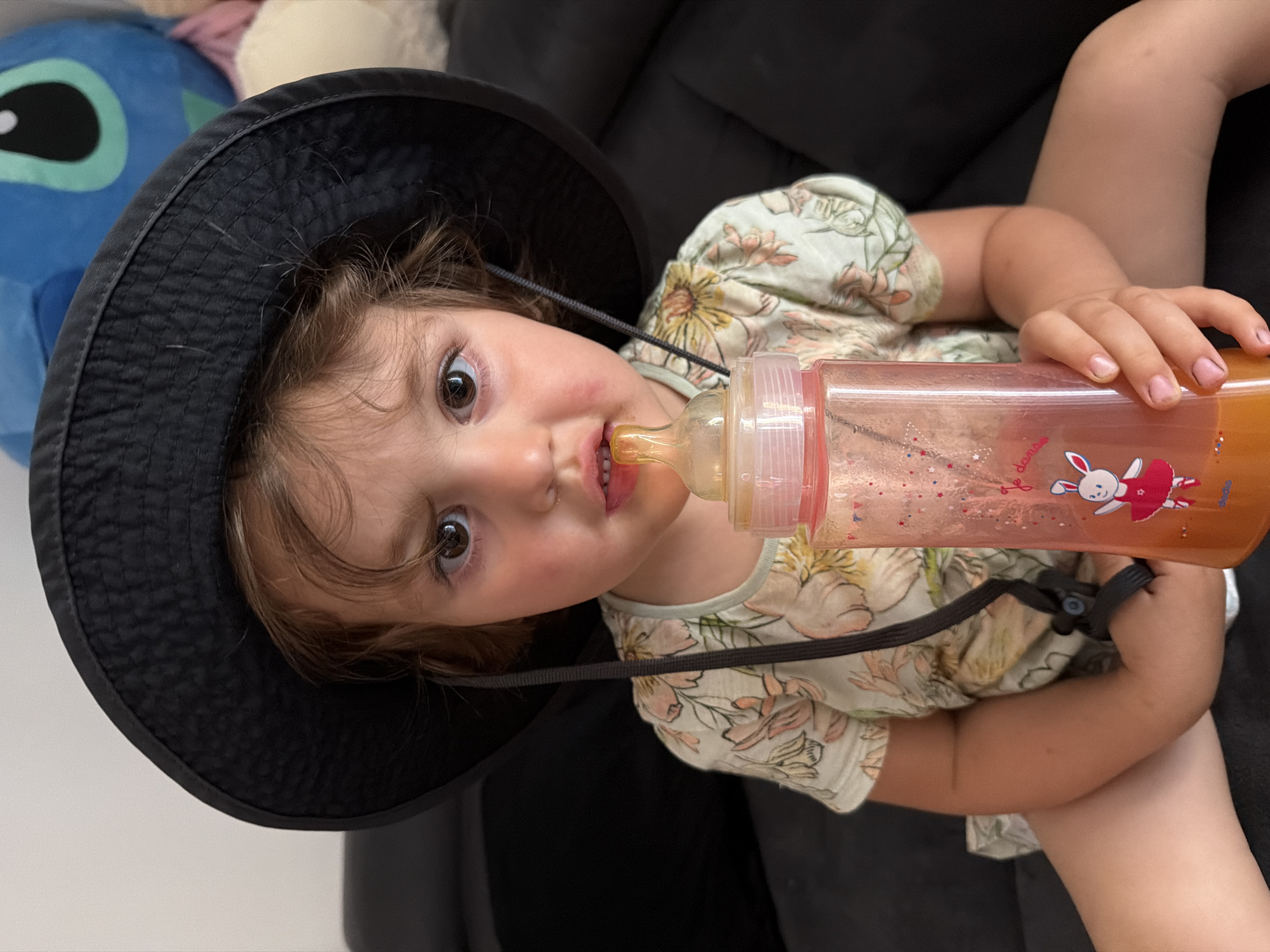 A young child wearing a black bucket hat and a floral print shirt is holding a pink baby bottle with orange liquid and a pacifier. The child is seated on a dark couch, with a blurred blue plush toy in the background. The image is taken indoors, likely in a residential setting, with no specific identifying features of Plovdiv, Bulgaria present.