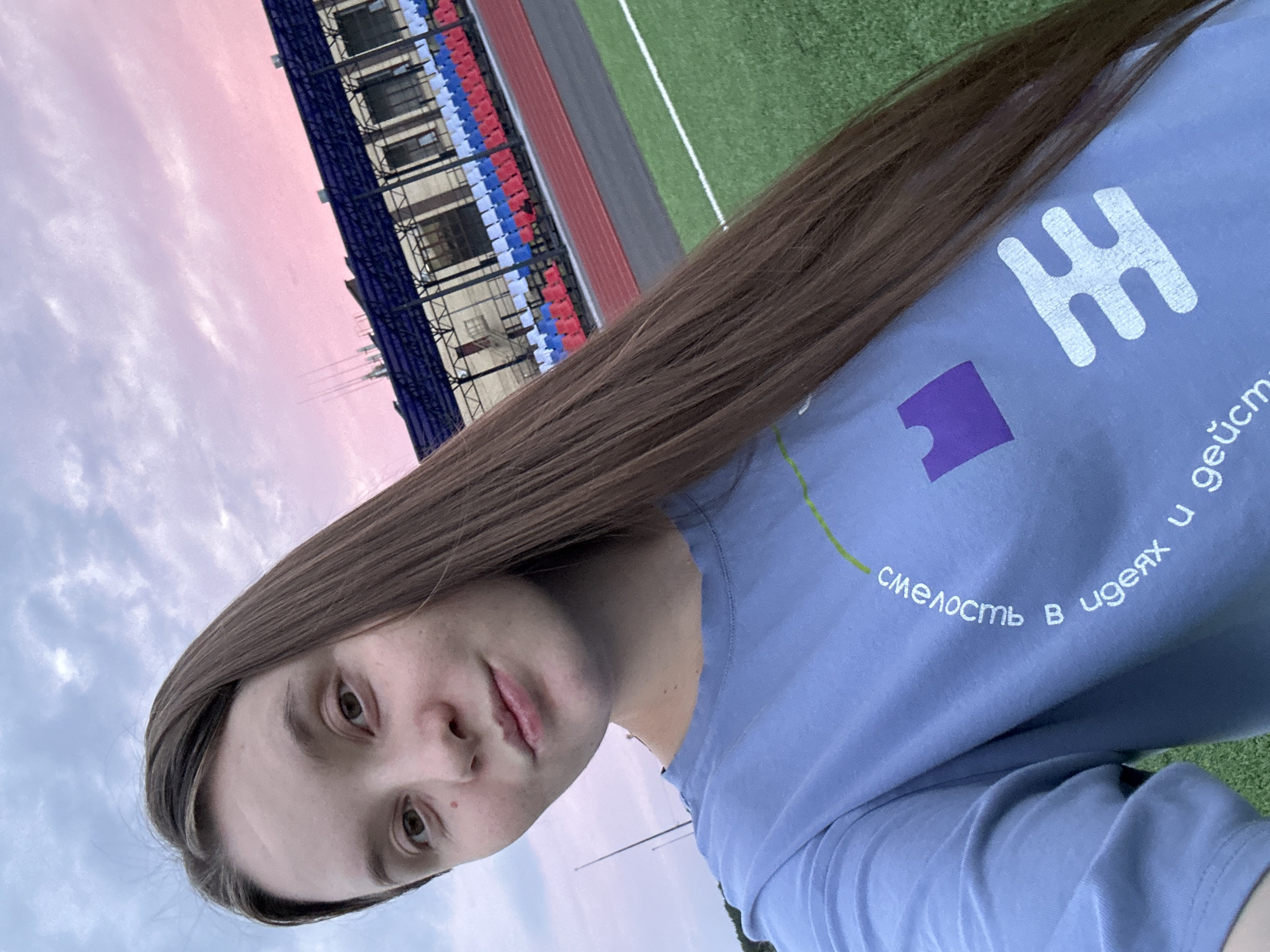 In Noviy, Russia, a young woman with long brown hair takes a selfie outdoors. She is wearing a light blue t-shirt with a graphic design and text in Russian that reads "Смелость в идеях и действиях" (Courage in ideas and actions). The background features a sports stadium with tiered seating in red, white, and blue, a running track, and a green field. The sky above is a twilight mix of gray clouds and soft pink hues.