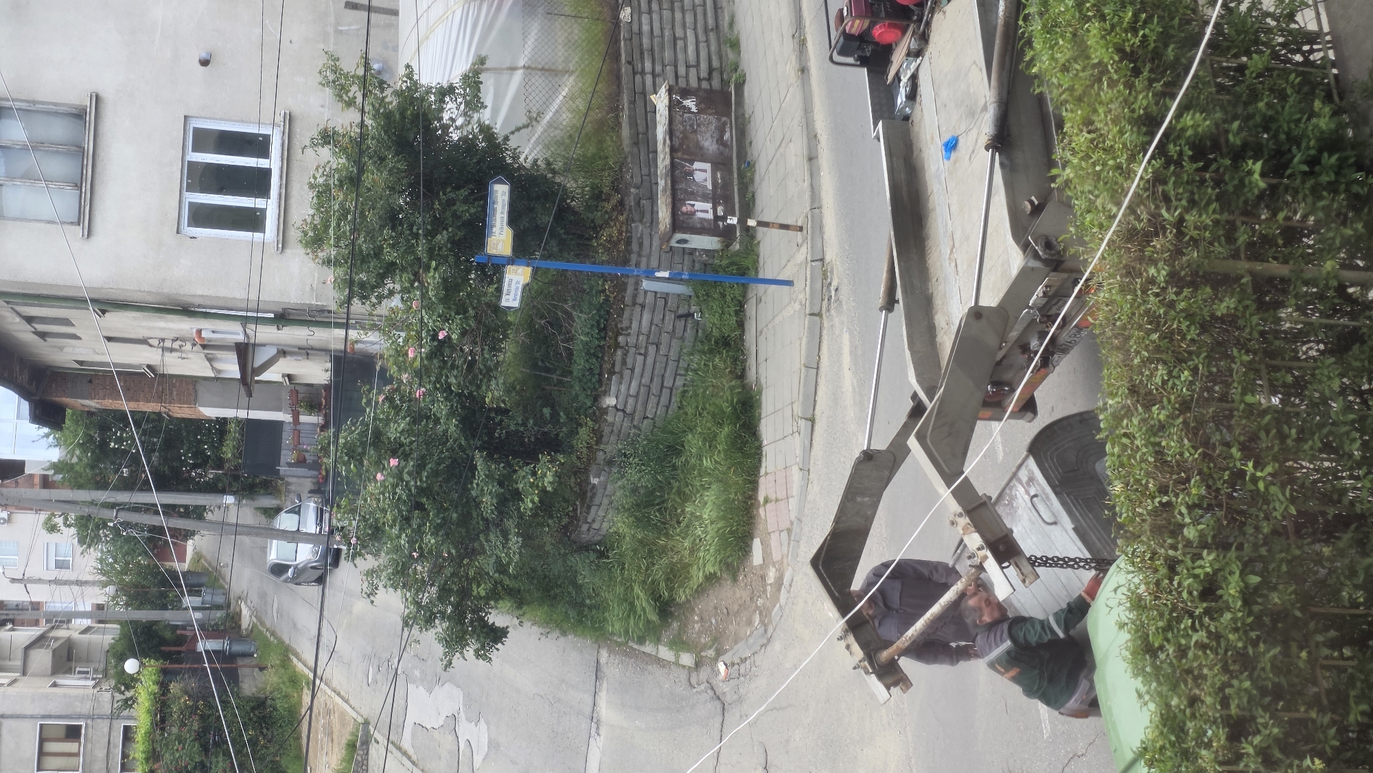 A street scene in Blagoevgrad, Bulgaria, shows a garbage truck with two men working to empty a green waste container. The truck's mechanical arms are lifting the container. A blue street sign indicates intersecting streets "Св. Мъченик Димитър Солунски" (St. Martyr Dimitar Solunski) and "Ж. Манушев" (J. Manushev). A gray building with white-framed windows is visible, along with a neighboring building with brick accents. The area is characterized by overgrown vegetation and utility poles with wires. A silver car is parked on the street in the distance.