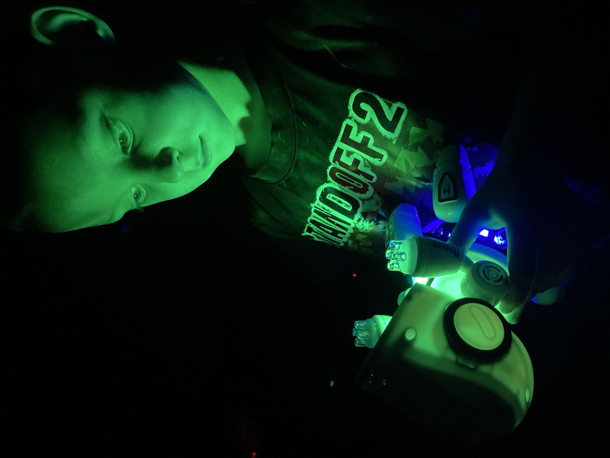 A person, likely a child or young adult, is captured in a dimly lit setting, with their face and upper body illuminated by a strong green light source. Their head is tilted, and their gaze is directed towards several glowing electronic devices. The green light casts a distinct hue on their skin and reflects off their eyes.
The person is wearing a black t-shirt with glowing green text, which clearly shows "WDOFF2". A prominent white, rounded electronic device, possibly a controller or toy, is in the foreground, featuring a large, glowing green circular button with a black rim. A hand, visible on the right, is holding another glowing device that emits both green and blue light from multiple small, bulb-like elements. Another smaller device, partially obscured, also shows a white "H" symbol and emits blue light.
The scene is almost entirely black, indicating a dark room or night-time environment where the glowing objects are the primary light sources. The activity appears to be interaction with or observation of these illuminated gadgets. The person's expression is one of focused attention or curiosity. There are no visible cues within the image itself to suggest the location of Noviy, Russia. The image is dominated by the stark contrast between deep shadows and the vibrant green and blue glows.