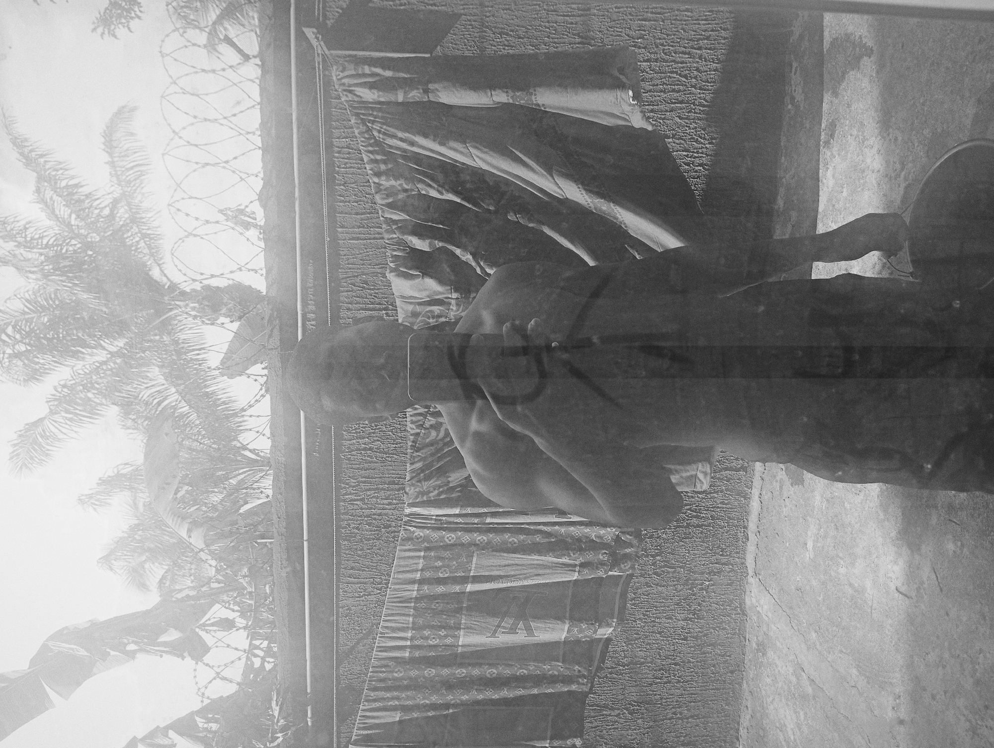 The photograph, taken in Nkpor, Nigeria, depicts a man standing behind a fence. The fence is topped with barbed wire and there are palm trees visible in the background. The man is holding clothing with a Louis Vuitton pattern in front of him. The surroundings appear to be outdoors with a concrete structure behind the man. The image is in black and white and seems to have been taken through a window or reflective surface, adding a layer of ambiguity to the scene.