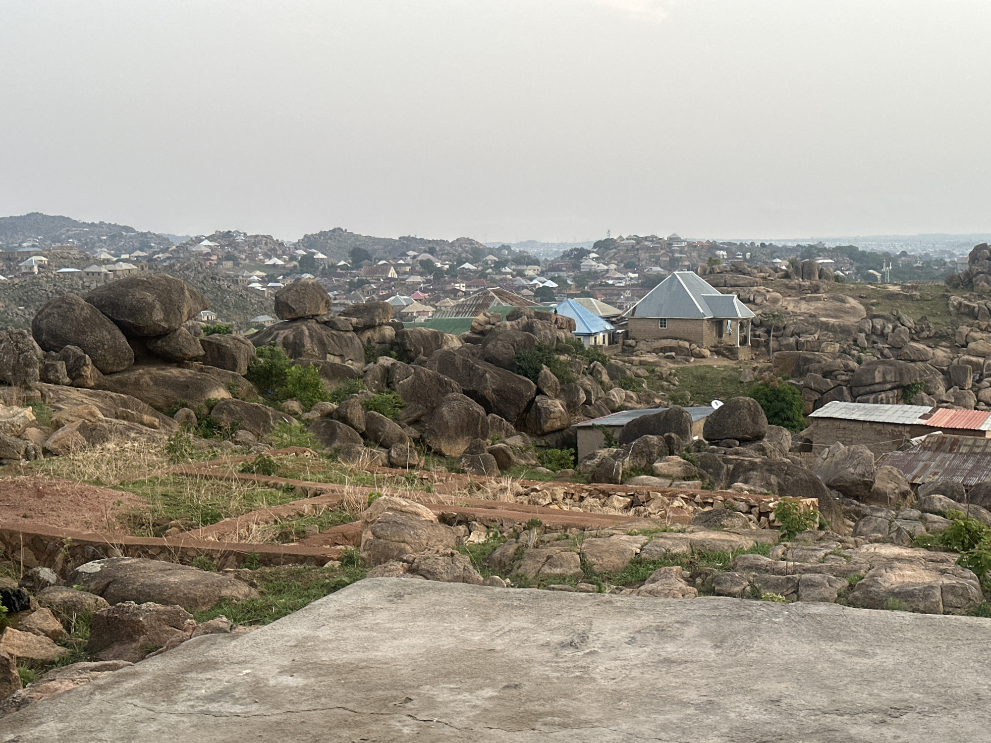 This wide-angle shot shows a portion of Jos, Nigeria, characterized by a landscape interwoven with large boulders and rock formations. Many structures are visible throughout the image. The buildings consist of houses with light-colored roofs, with a few having blue or red roofing material. In the foreground, there's a concrete surface with rocks. Beyond that, there is a dirt path. The setting is overcast, contributing to a muted color palette. The image's composition places the viewer above the scene, looking out towards the horizon.