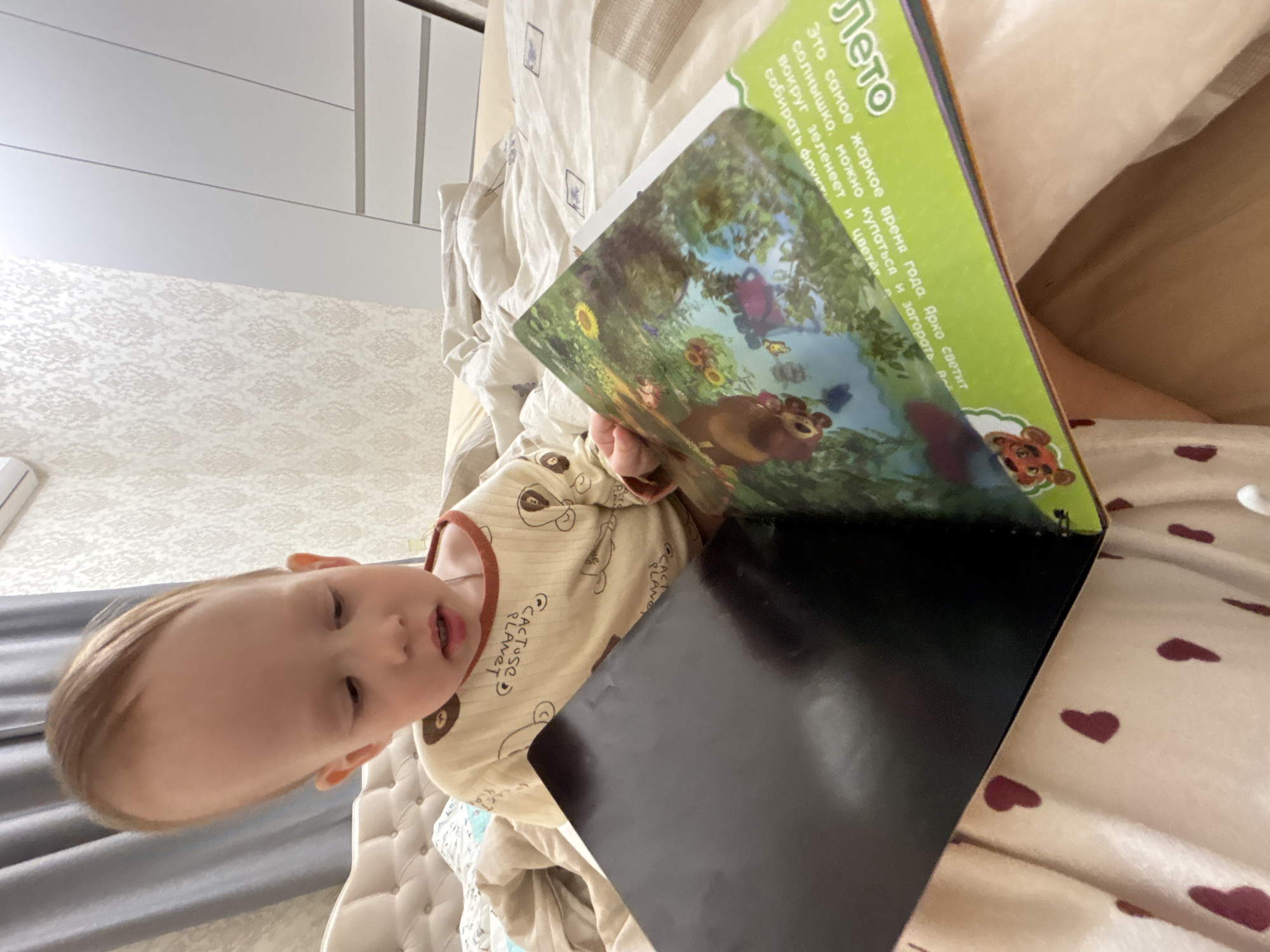 A child is lying on a bed, appearing to read a book with a green cover and illustrations of a bear and greenery. The cover text is in Russian. The child is wearing a beige long-sleeved shirt with cartoon images. The bed has patterned sheets and cushions. The location is the city of Ферма, Russia.