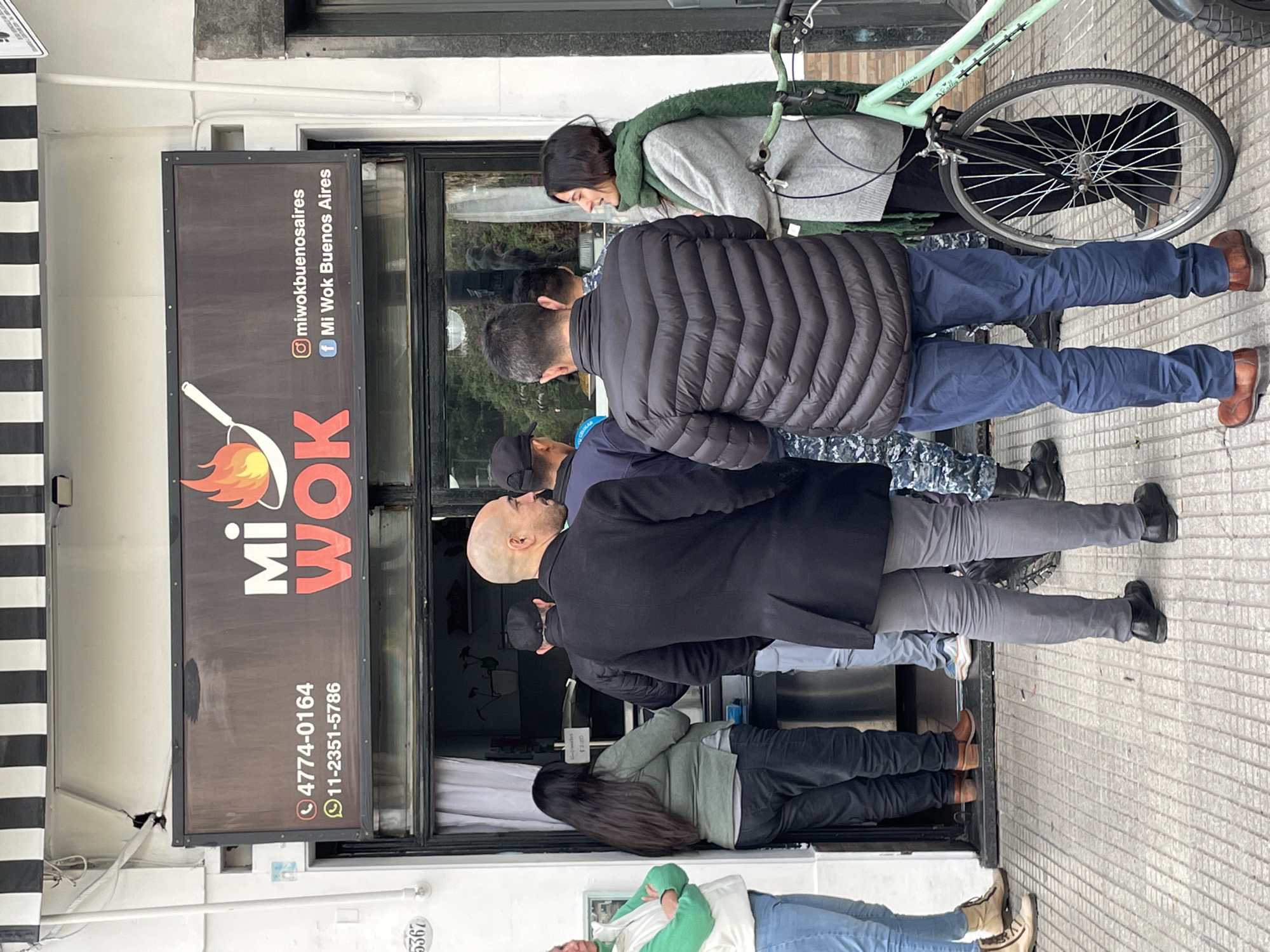The image depicts an urban street scene in Buenos Aires, Argentina, outside a restaurant named "Mi Wok." Several people are standing in a queue or gathered near the entrance of the establishment.
In the foreground, a person with a bald head, wearing a black coat and grey pants, stands facing right. Behind them, another individual wears a black puffer jacket, blue pants, and brown shoes. A woman with dark hair, dressed in a grey sweatshirt and a green scarf, is visible further back, standing near a mint green bicycle. Other people, including one in a sage green jacket and dark pants, and another in a green hoodie and light jeans, are partially visible in the line or inside the store.
A prominent dark sign for "Mi Wok" is affixed to the storefront, featuring a logo of a flaming wok. The sign clearly displays social media handles: Instagram "@miwokbuenosaires" and Facebook "Mi Wok Buenos Aires." Two phone numbers are also visible: "4774-0164" and "11-2351-5786." Inside the window, a small, partially obscured sign shows "2.000," likely indicating a price. The building features a light-colored facade and a black and white striped awning. The ground is tiled pavement. The overcast lighting suggests a daytime setting with cooler weather, matching the warm clothing worn by the individuals. A building number "5362" is also visible on a fixture.