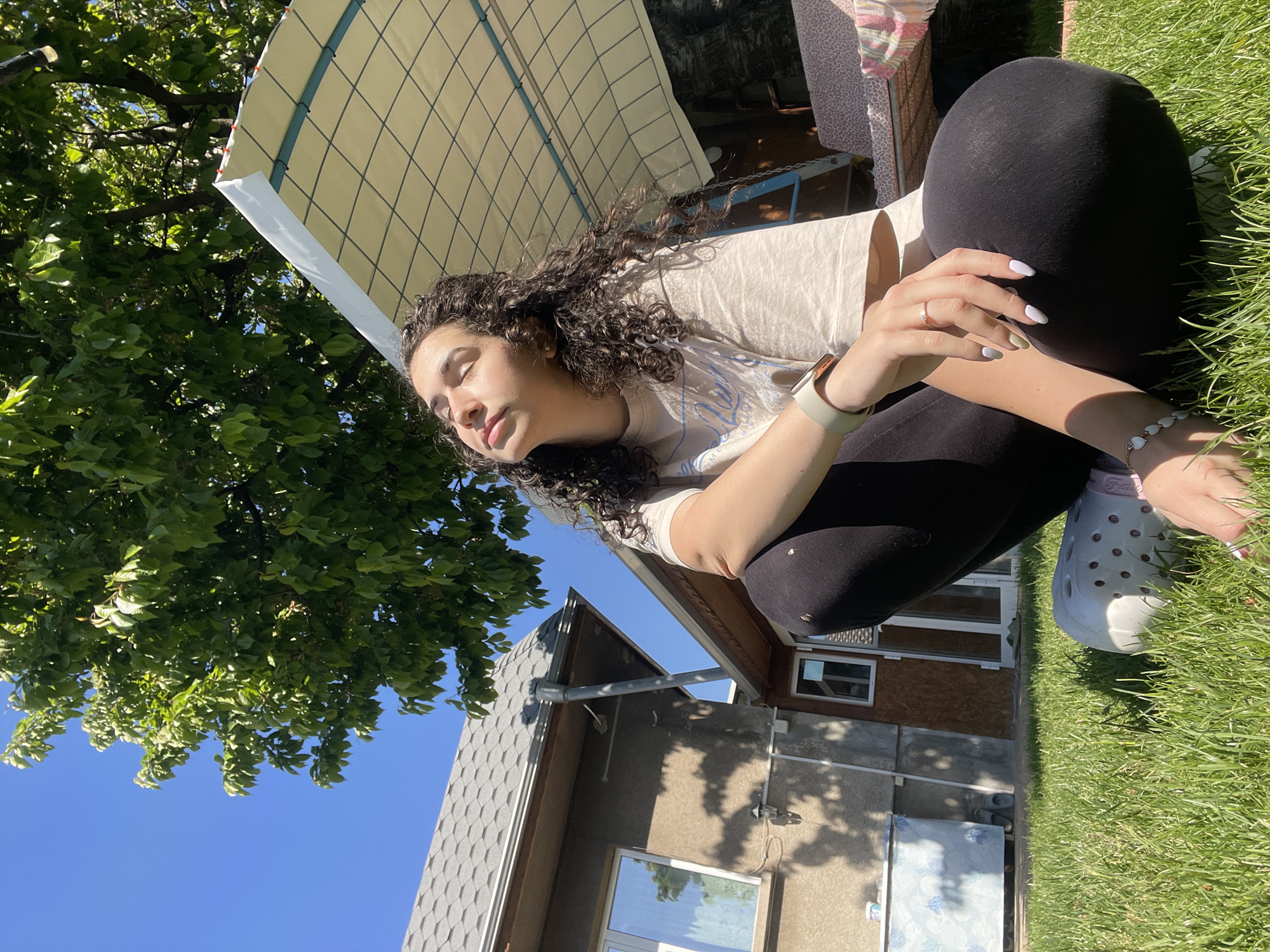 A young woman with curly hair is sitting on green grass outdoors, possibly in a yard or park. She's wearing black leggings, a light-colored t-shirt, and white Crocs. Her eyes are closed, and she appears to be relaxing in the sunlight. She has a watch on her left wrist, a ring on her finger, and a bracelet on her right wrist.
In the background, there's a house with a gray roof and brown siding. A tree with green leaves is visible on the left side of the image, and a canopy structure is overhead, partially obscuring the sky. The sky is a clear, bright blue.
Without specific landmarks, providing an exact location in Sofia, Bulgaria, is impossible. However, the scene suggests a residential area with yards and houses, possibly on the outskirts of the city.