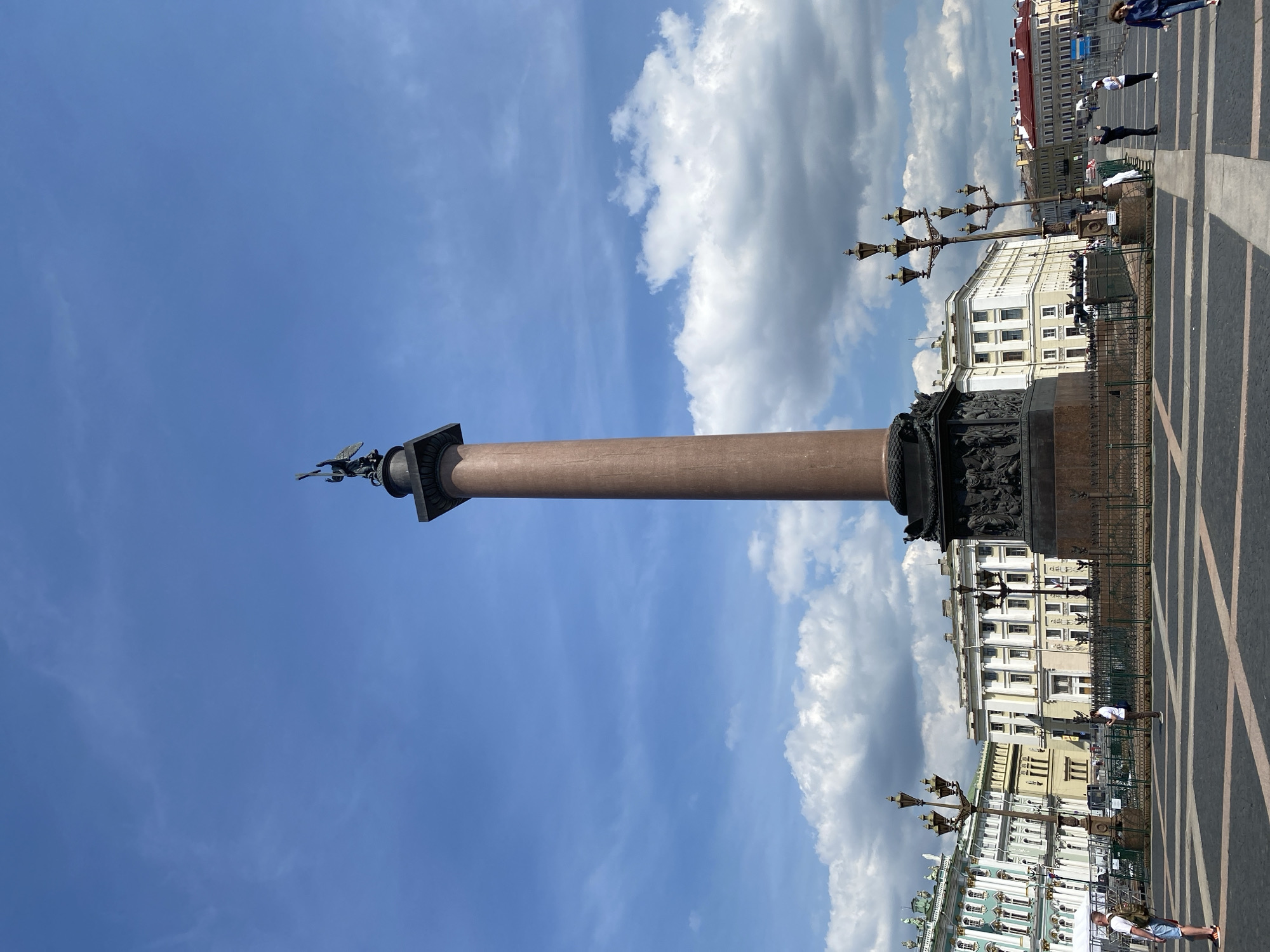 The image shows a low-angle, full-shot view of the Alexander Column in Palace Square, Saint Petersburg, Russia. The column, made of red granite, stands tall against a bright blue sky with scattered white clouds. Atop the column is a bronze statue of the Angel of Peace. The base of the column is adorned with intricate bronze reliefs depicting scenes from Russian military history. Surrounding the column is a black metal fence. In the background, grand 18th and 19th-century buildings with classical architecture line the square, including the General Staff Building and the Winter Palace. Several lampposts with ornate designs are visible throughout the square. A few individuals are scattered across the paved square, some walking, others standing and observing the monument. The ground is paved with dark cobblestones and lighter stone inlays forming geometric patterns.