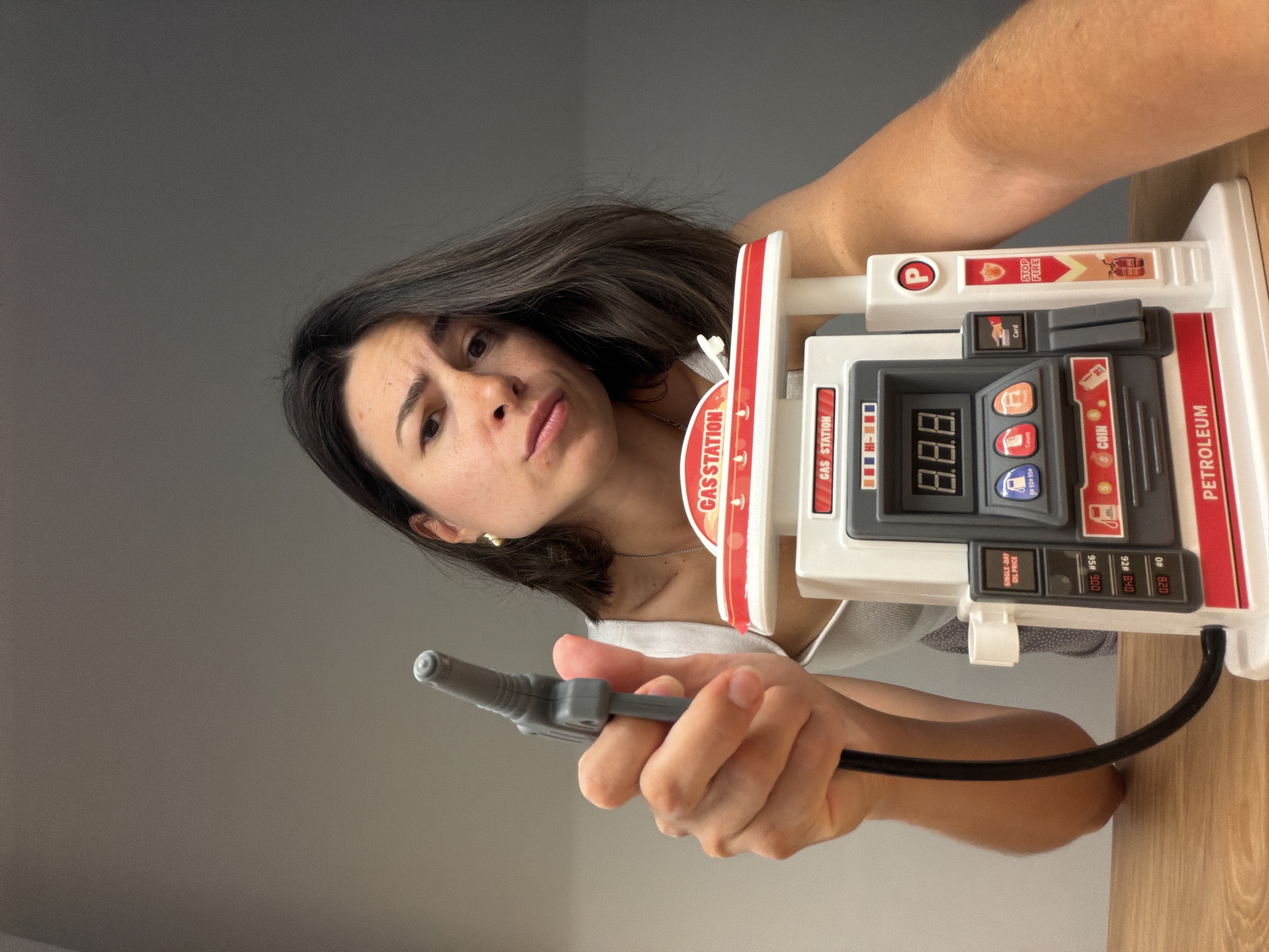 A woman is holding a toy gas pump, as if she is pumping gas. The toy gas pump is white, red, and grey. The woman has dark hair and is wearing a white top. She has a serious expression on her face. The background is a plain grey wall and a light brown wooden surface. There is no indication of the location being in Budva, Montenegro.