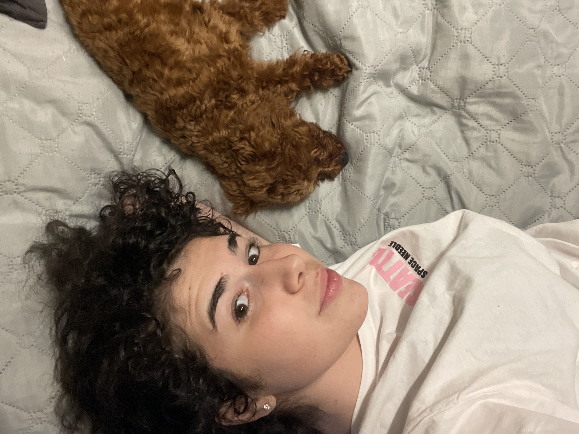 A woman with curly dark hair is lying on a gray quilted bedspread, looking towards the camera. A brown poodle is lying on the bedspread above her head. The woman is wearing a white t-shirt with "SPACE NEEDLE" printed on it. The scene is inside, likely a bedroom. The image does not provide any information related to Sofia, Bulgaria.