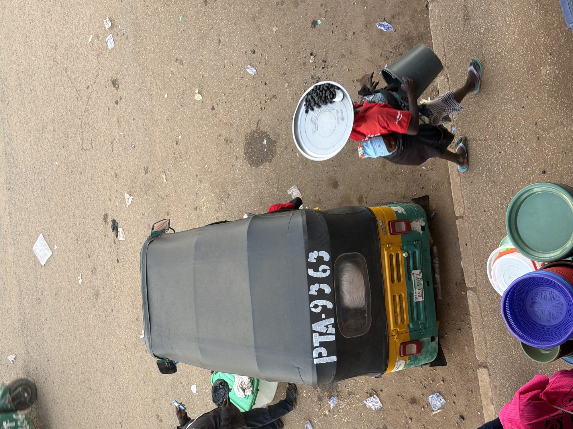 This image depicts a street scene in Bukuru, Nigeria. A tuk-tuk, with license plate RSA-50YD and registration number PTA-9363, is parked on a dusty road. A person stands next to the tuk-tuk holding a mobile phone and a green container. Further down the road, a woman is walking while carrying a child on her back. She is also carrying a large black bucket in her right hand and a silver tray with a pile of dark berries on her head. In the foreground, several colorful plastic baskets are stacked. The ground is covered in debris, suggesting a busy and potentially unkempt urban environment.