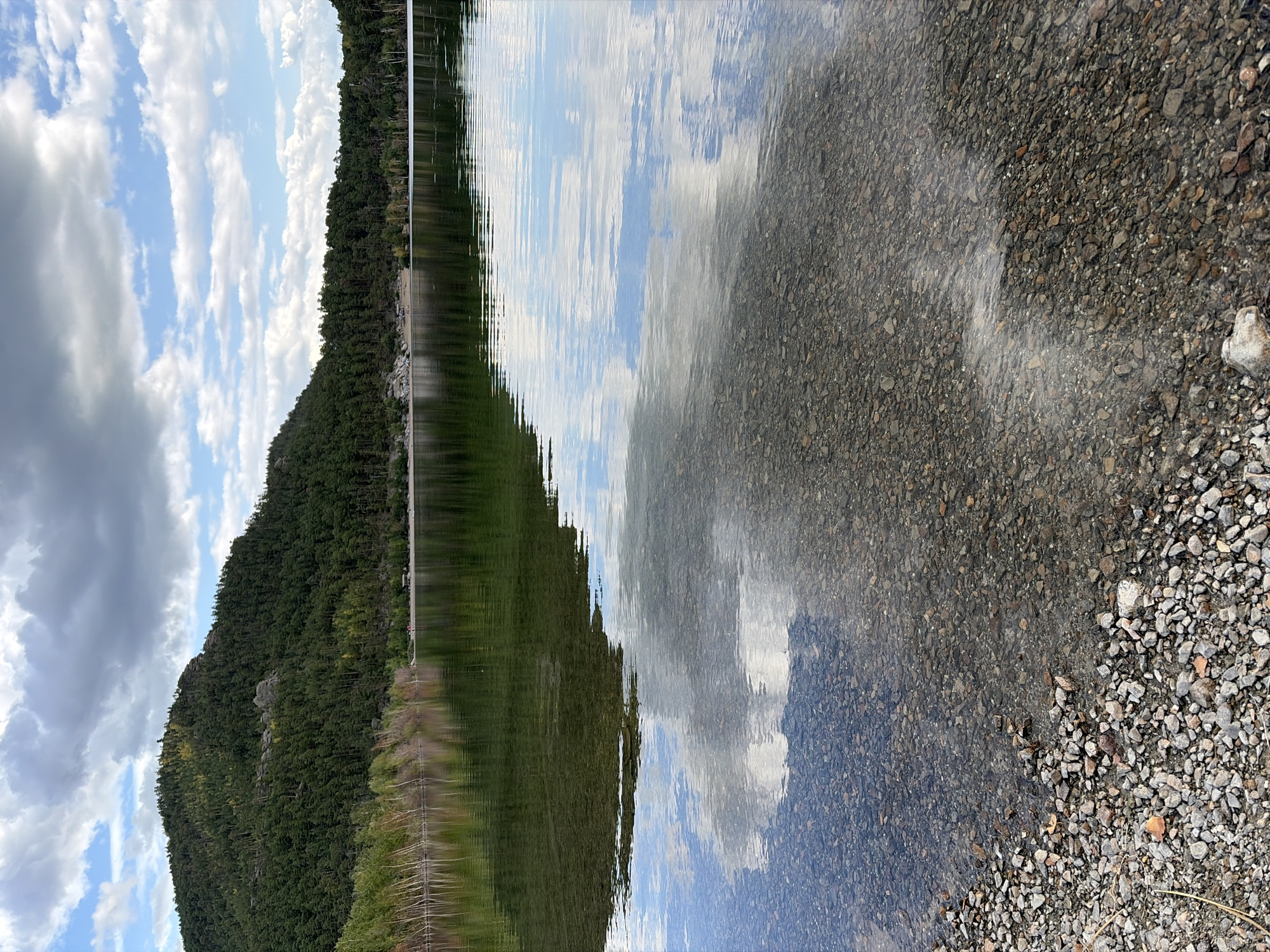 The image depicts a serene natural landscape at a lake in Зелёный Бор ауылдық округі, Kazakhstan. In the foreground, a rocky shore is visible, with the clear water revealing the pebbled lakebed. The water's surface reflects the sky, showing white clouds against a blue expanse. Beyond the water, a tree-lined shore stretches across the midground. The background is dominated by a lush, tree-covered hillside or mountain. Several small figures, likely people, are discernible on the distant shore, near a structure that could be a small building or shelter. The overall scene suggests a tranquil, outdoor environment, possibly a recreational area.