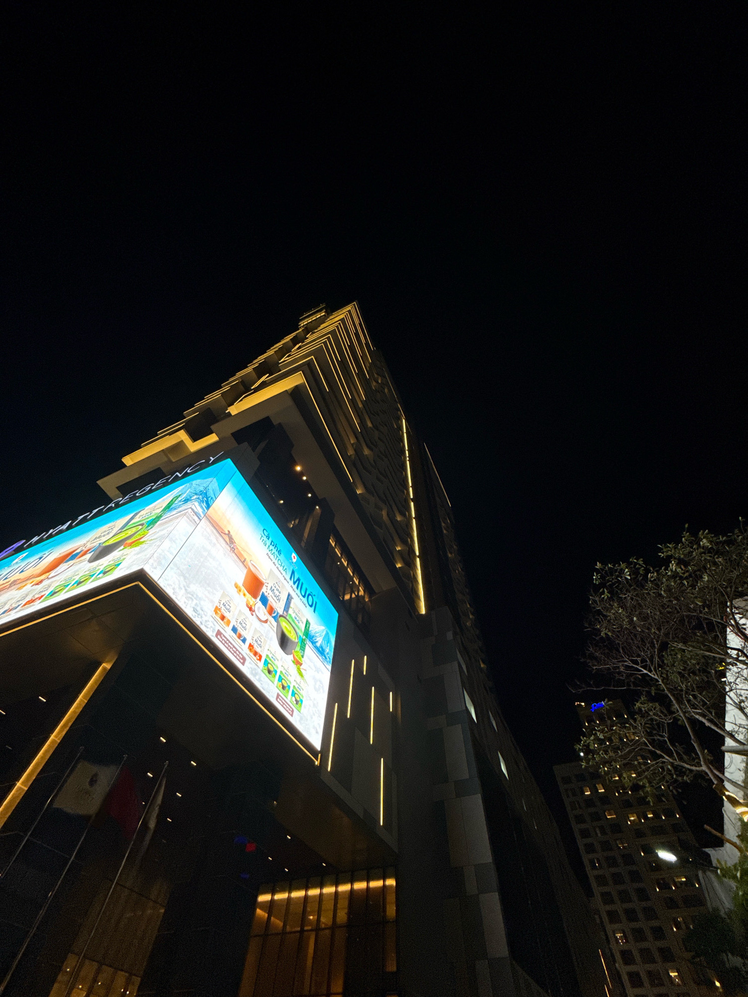 This image shows a low-angle, night-time shot of a modern skyscraper in Nha Trang, Vietnam. The building is illuminated with warm, yellow lights accentuating its geometric facade. A large, brightly lit digital billboard wraps around a corner of the building, displaying an advertisement for matcha tea with images of green tea products and scenic landscapes. Below the billboard, two flags are visible on flagpoles, one appearing to be the Mexican flag. To the right, another skyscraper with illuminated windows can be seen in the distance, along with a silhouetted tree. The sky is completely dark, indicating the time is late at night.