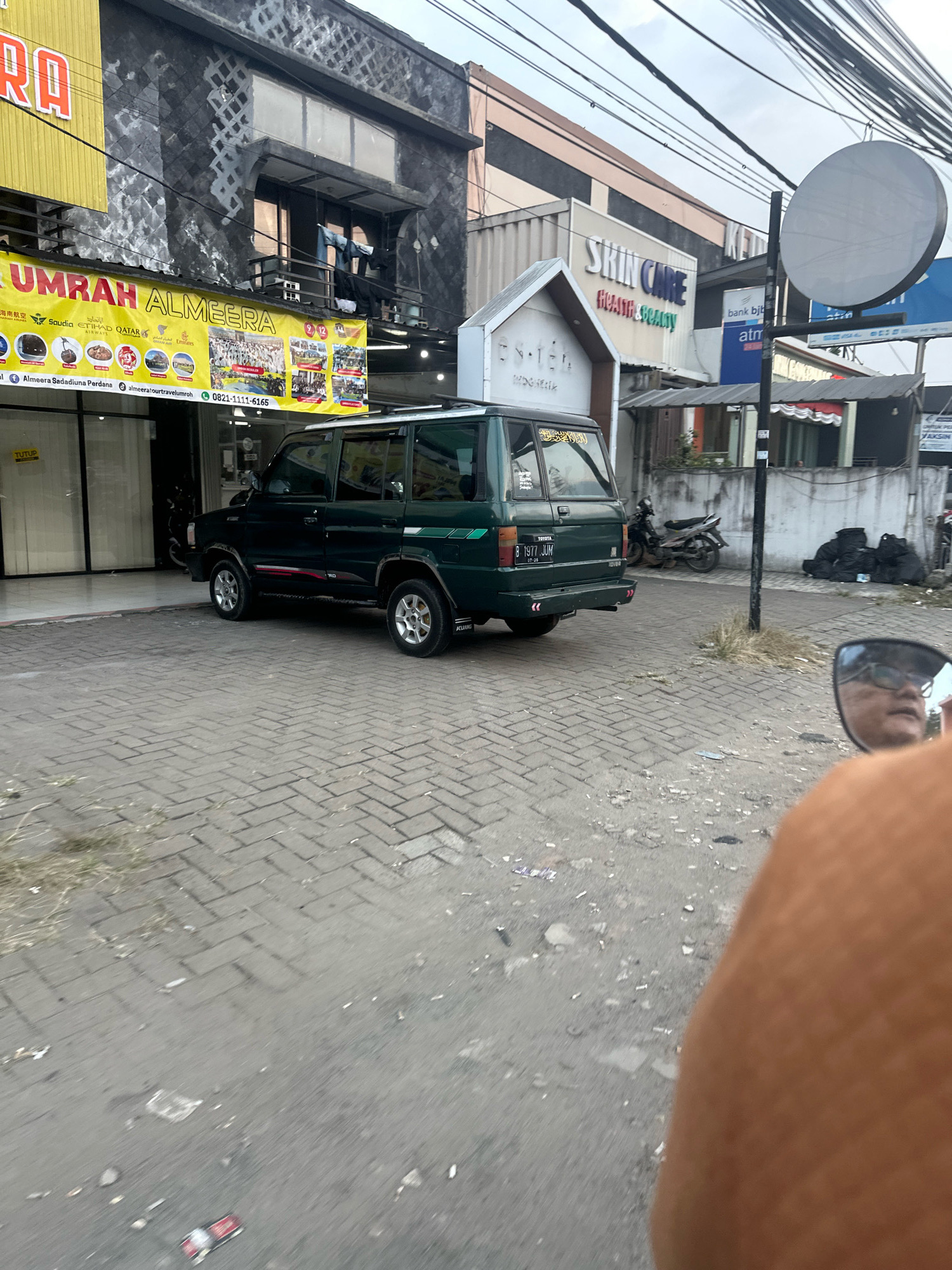 The image shows a street scene in front of commercial buildings. A dark green Toyota Kijang is parked on a paved area. A yellow banner with the text "UMRAH ALMEERA" and various logos is displayed on the building to the left. To the right, signs for "SKIN CARE" and "bank bjb" are visible. The street is paved with interlocking bricks. In the foreground, a person's shoulder and arm are visible, suggesting the photo was taken from a moving vehicle. The environment appears to be an urban area, possibly in Saruni, Indonesia, given the Indonesian license plate on the car and the context of the provided crops. There are power lines and a traffic light visible in the background. Bags of trash are piled on the right side of the street. A motorcycle is parked behind the car.