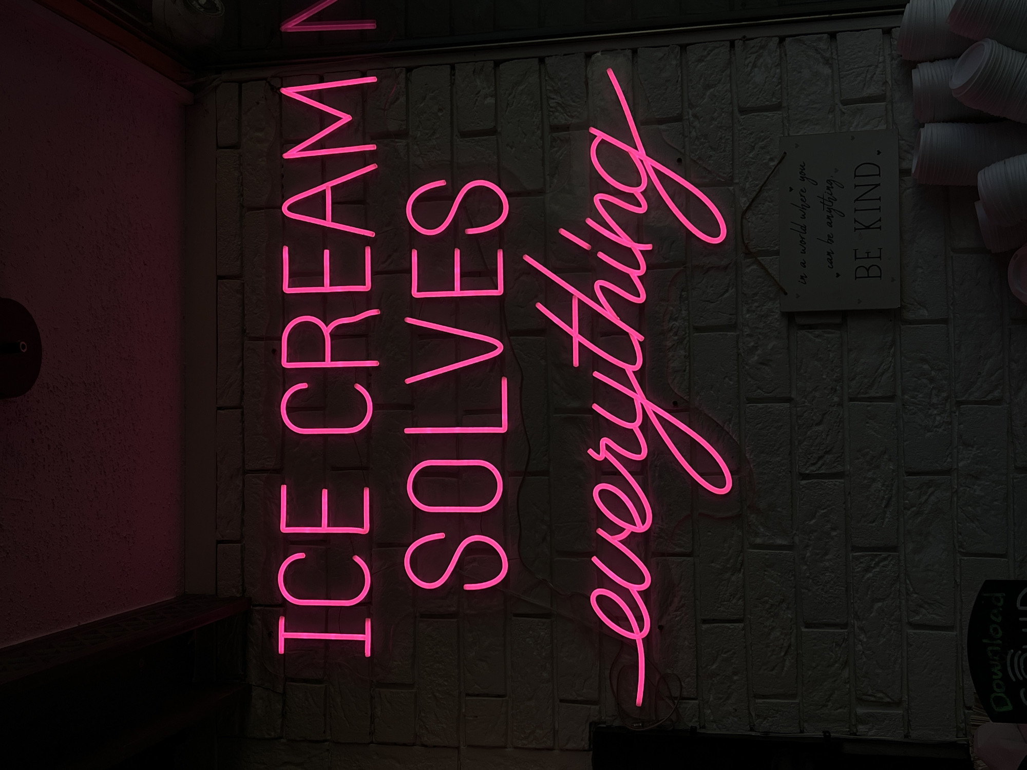A neon pink sign displaying the text "ICE CREAM SOLVES everything" is mounted on a white brick wall. Below the neon sign, a small wooden sign reads "in a world where you can be anything. BE KIND." In the bottom right of the image, stacks of white paper cups are visible. The scene appears to be indoors, possibly in an ice cream shop or similar establishment. There are no people visible in the image. The lighting is dim, with the neon sign being the primary light source. The provided information about the location being in Donegal, Ireland cannot be confirmed from the visual evidence in the image.