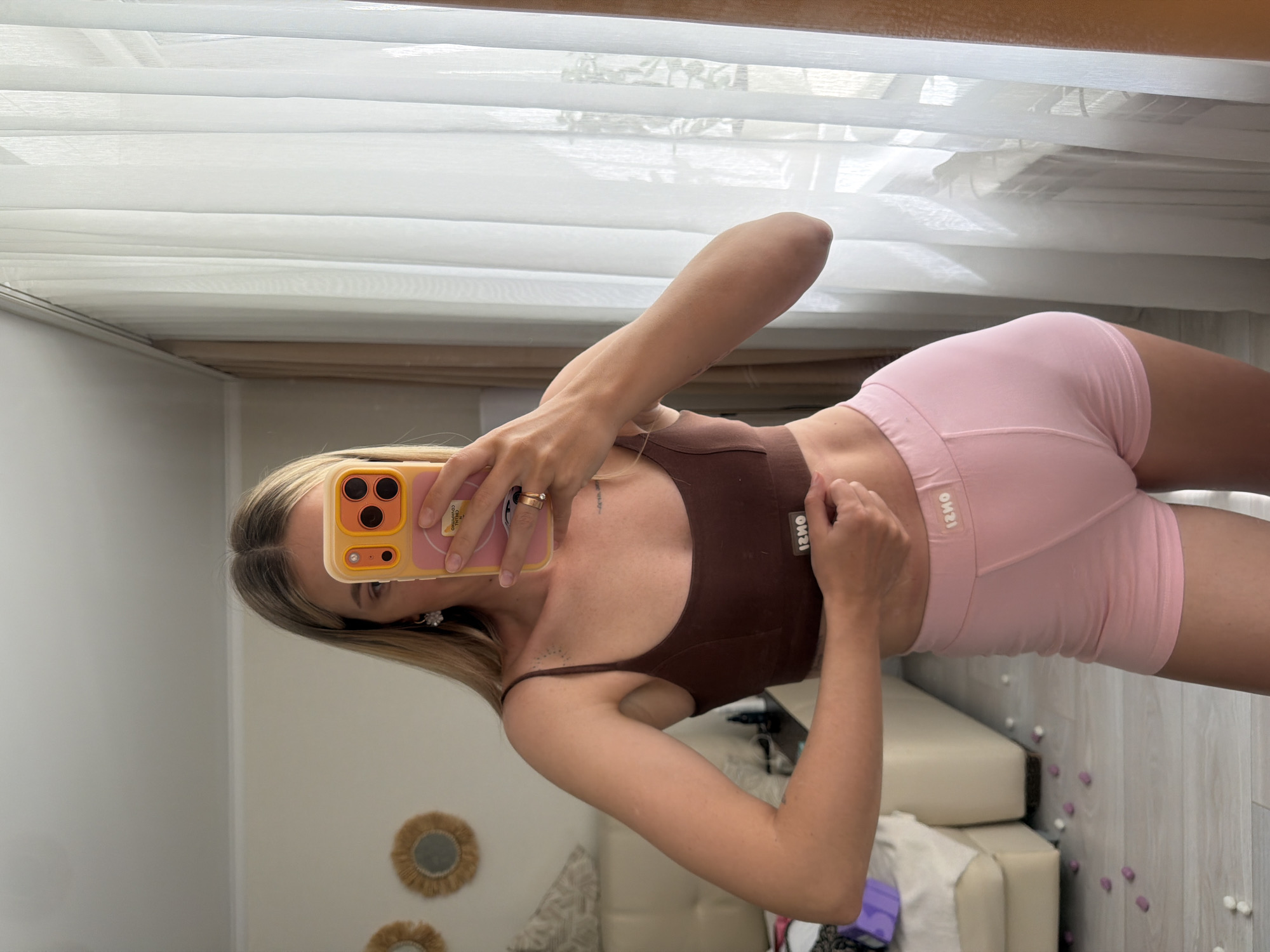 A blonde woman is taking a mirror selfie in a bedroom. She is wearing a brown sports bra and pink shorts. She is holding a smartphone in her right hand and has her left hand on her stomach. The room is sparsely decorated with two circular wall hangings on the left and sheer curtains on the right. A bed and a couch are visible in the background. The floor is light-colored wood, with scattered small white and pink objects. The overall scene suggests a casual, personal moment. This image does not provide enough information to determine the location in Semiluki, Russia.