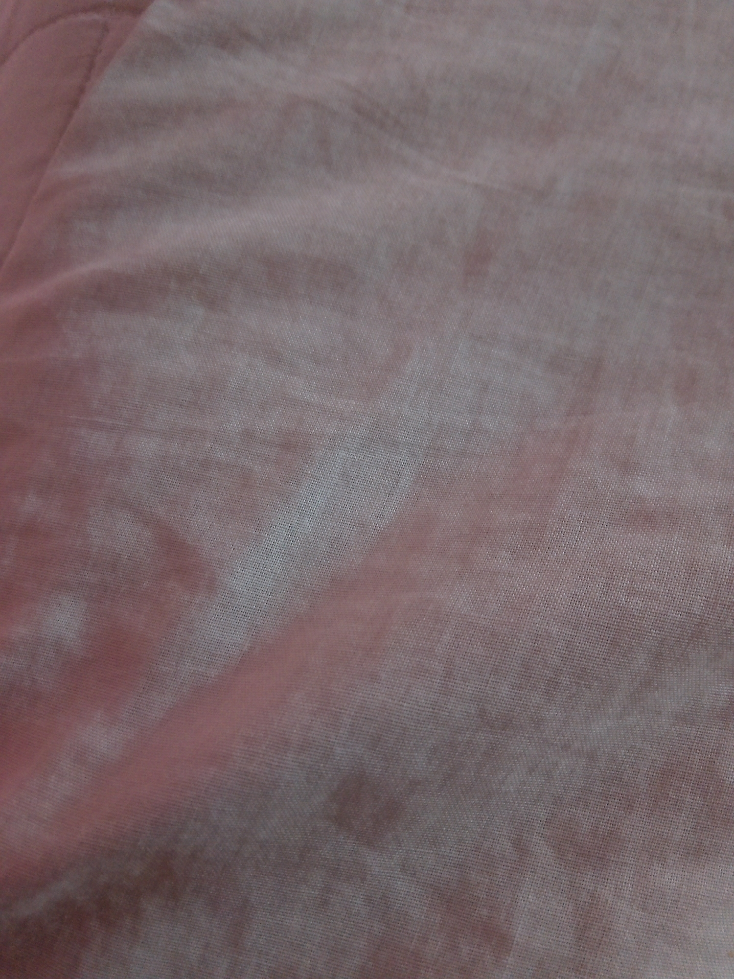The image displays a close-up view of a textured, light pink fabric. The fabric appears to be wrinkled and creased, with subtle variations in shading and tone, suggesting it might be a textile like cotton or linen. There are no discernible objects, people, actions, or interactions visible within the frame. The scene is a plain, indoor shot focusing solely on the material's texture and color. There is no indication of its location within Mansehra, Pakistan.