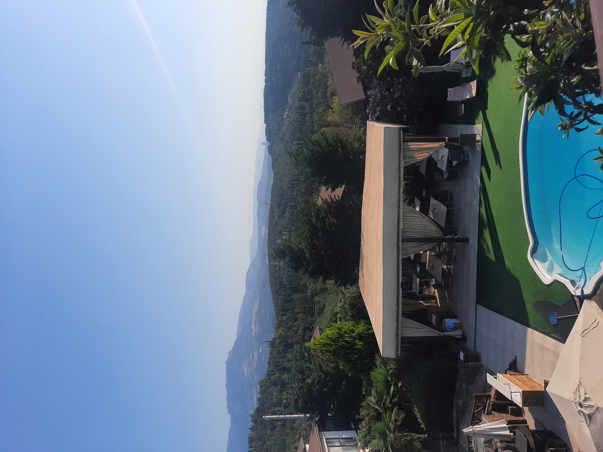 A high-angle, wide shot captures a resort area with a swimming pool and an outdoor dining pavilion, set against a backdrop of lush, green hills and distant mountains under a clear blue sky. In the foreground, a bright blue swimming pool with a curly blue hose is partially visible. To the left of the pool, an area covered with artificial turf hosts an outdoor dining pavilion with several tables and chairs, all under a beige canopy. Beyond the pavilion, a building with a brown roof is partially visible on the left. The middle ground is dominated by dense green trees and cypress trees covering the rolling hills, with some hints of buildings nestled within the foliage. In the background, layered mountain ranges recede into the distance, their peaks softened by haze. The overall scene is peaceful and serene, suggesting a warm, sunny day. There are no people or animals visible in the frame.