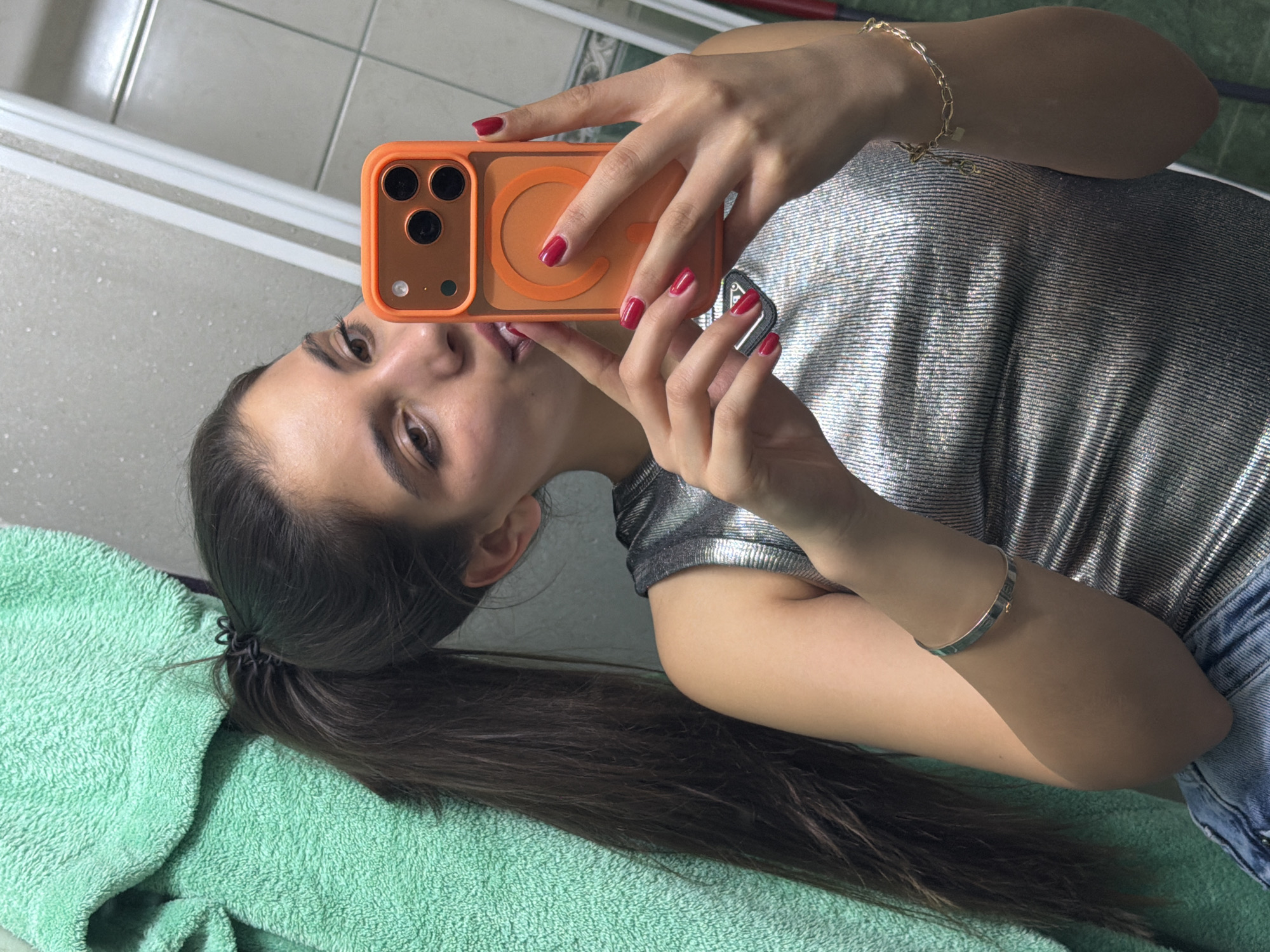 A young woman is taking a selfie in a bathroom. She is holding an orange iPhone with red nail polish on her fingers. The woman is wearing a silver shirt and a ponytail. A green towel is visible in the bottom left corner. The background shows a white tiled wall and a shower door. The location is likely Rudartsi, Bulgaria based on the user's information.