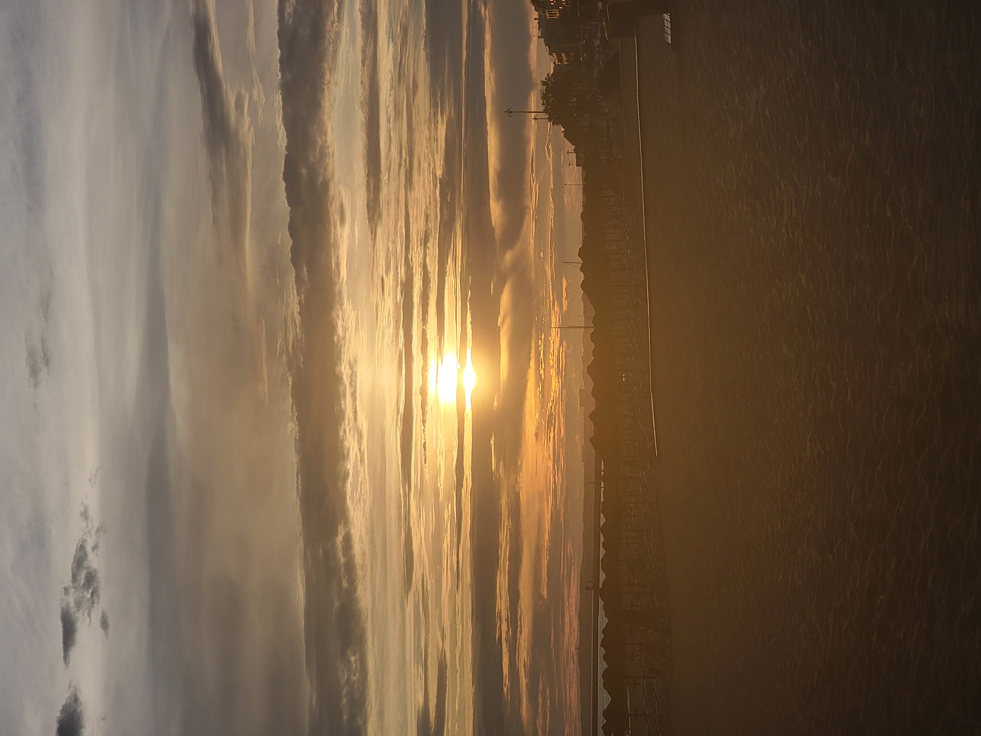 The image depicts a beach scene at sunset in Quarteira, Portugal. The sky is filled with dramatic clouds illuminated by the setting sun, which is visible as a bright orb near the horizon. The warm hues of orange and yellow cast a golden glow over the scene. In the foreground, the sand of the beach is textured, showing subtle ripples. Beyond the sand, rows of empty beach umbrellas and loungers are set up, suggesting a quiet moment before or after a day at the beach. In the distance, along the coastline, buildings and structures of the town are visible, silhouetted against the colorful sky. The overall atmosphere is serene and peaceful.