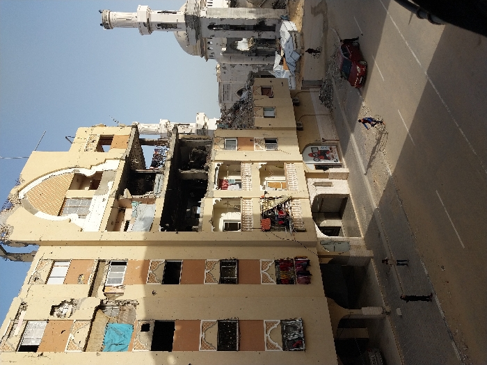 The image captures an urban street scene in Khan Yunis, Palestinian Territory, displaying extensive damage to buildings under a clear, bright sky. The prominent structure in the left and central foreground is a multi-story building, primarily beige with orange-brown sections and decorative arched motifs, exhibiting severe structural damage. Multiple floors have large sections of exterior walls missing, revealing interiors, exposed rebar, and debris. Windows are broken or absent, and several balconies show hanging laundry, including blue, red, and pink items. The ground floor features arched entryways, and a visible storefront displays a "SQUID GAME" poster. To the right and rear of this building, a mosque with a distinctive minaret and a dome is visible; it also appears damaged or to be under construction/deconstruction, with visible rebar at the top of the minaret and rubble accumulated around its base. Piles of construction debris and structures covered with white tarpaulins are situated between the large building and the mosque. A paved asphalt road with white lane markings extends across the lower portion of the frame. On the left sidewalk, three individuals are standing. In the road, one person rides a bicycle to the right. A red sedan is parked on the right side of the road with its driver's side door open, and one person stands next to it. A silver car is partially visible parked under an archway on the left sidewalk. Shadows are cast to the left, indicating the sun's position.