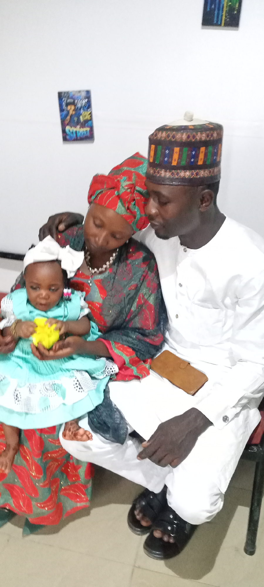 A photograph shows a family consisting of a man, woman, and baby sitting together in an indoor setting. The woman, wearing a red and green patterned headwrap and dress, holds the baby in her lap. The baby is wearing a light blue dress with a white bow in her hair and is holding a yellow object. The man is sitting next to the woman, dressed in a white shirt and a patterned hat. He has a brown wallet on his lap and is wearing black sandals. The background consists of a white wall with two small, framed pictures hanging on it.