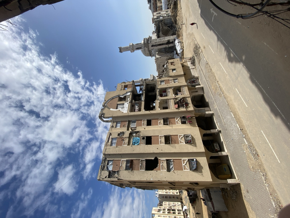 This image depicts a heavily damaged urban streetscape in Khan Yunis, Palestinian Territory, under a partially cloudy blue sky. In the foreground, a paved two-lane road with a double white centerline runs horizontally, bordered by a grey sidewalk. Shadows are cast across the street, indicating bright sunlight.
The central feature is a seven-to-eight-story light brown residential building with darker brown accent panels and decorative white window surrounds. The building exhibits extensive structural damage, including numerous impact holes, shattered or missing windows, and significant collapse on its upper right facade, exposing internal concrete and twisted rebar. Balconies with debris or fabric are visible. On the ground floor, an arched arcade provides covered parking, sheltering at least two vehicles, one yellow and one silver. Debris is accumulated at the base of the building.
To the right of the central building, a severely damaged mosque is visible, with its minaret still partially standing but exhibiting significant structural compromise. The main body of the mosque is largely reduced to rubble and twisted metal. Large piles of dirt and construction debris surround the mosque structure. A solitary individual wearing a red top and dark trousers walks across the dirt ground between the road and the mosque's remains, moving towards the right.
In the mid-ground to the left, several other multi-story residential buildings, similar in architectural style but appearing less severely damaged, line a dirt-covered area. Two small figures are visible walking on a path within this area. Overhead, thin electrical wires or communication lines traverse the sky in the upper left quadrant. The overall environment suggests significant destruction and displacement within a residential and religious area.