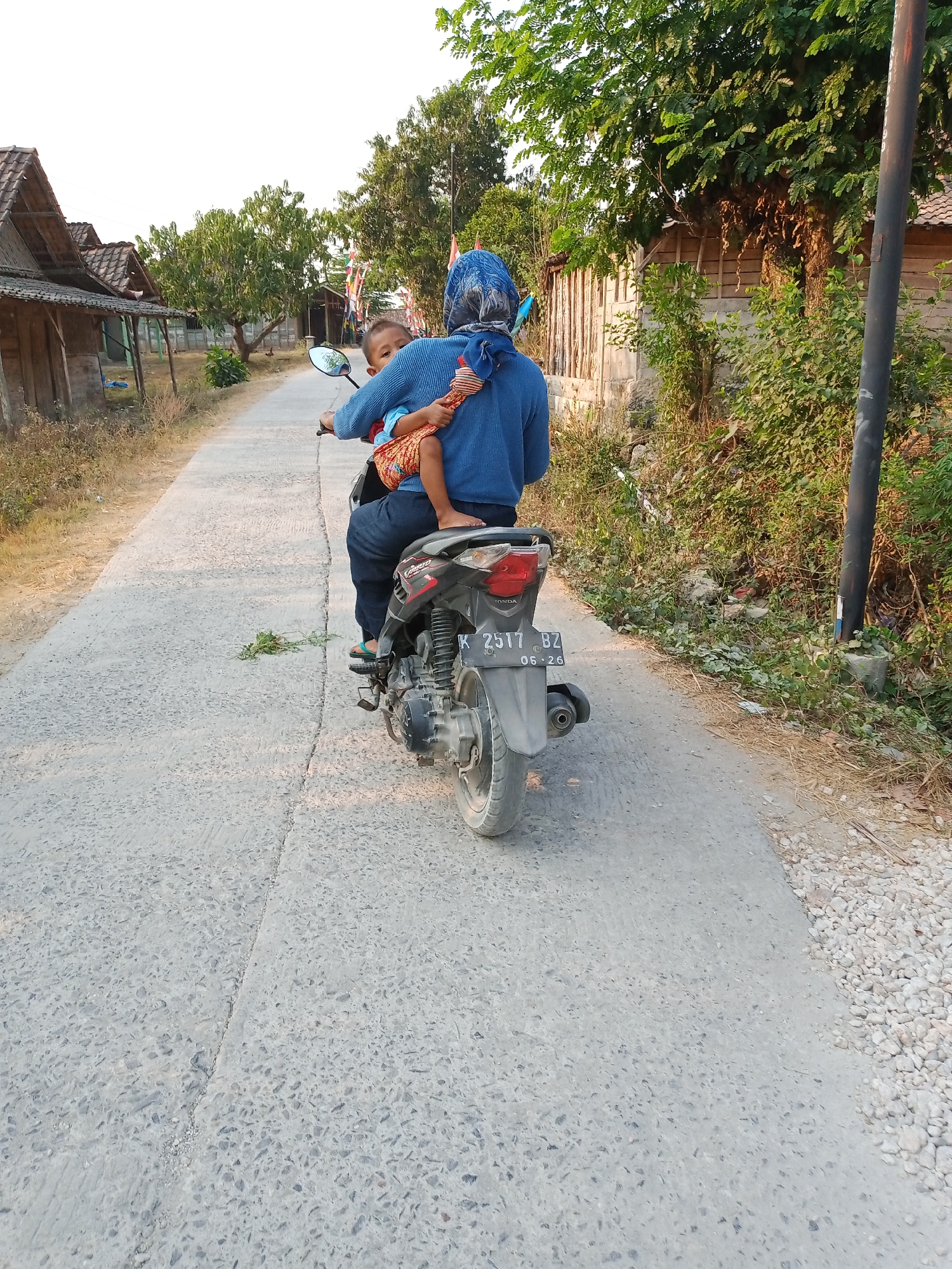 A woman wearing a blue headscarf and a blue sweater is riding a motorcycle with a young child sitting in front of her. The child is wearing an orange and white striped shirt and an orange and white patterned sarong. They are on a paved road in a rural area of Rowobungkul, Indonesia. The road is bordered by dry grass and vegetation. In the background, there are several small wooden houses with thatched roofs, some trees, and a few colorful flags. The sky is bright and clear. The motorcycle has a license plate that reads "K 2517 BZ".