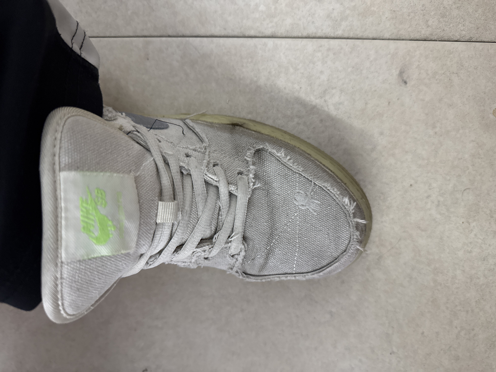An overhead shot captures a single, well-worn, light grey canvas shoe resting on a light-colored tiled floor in Yerevan, Armenia. The shoe, likely a sneaker, shows significant signs of use, including frayed edges around the laces and toe area, and some discoloration on the cream-colored rubber sole. White shoelaces are tied loosely. A small portion of a black garment, possibly pants or shorts, is visible to the left, indicating the shoe is being worn.
A prominent light green tag is affixed to the tongue of the shoe, clearly displaying the text "NIKE SB" with the iconic Nike swoosh logo above "SB." The upper of the shoe features stitched details, and some areas of the canvas appear textured or slightly roughened from wear. The tiled floor has faint grout lines and scattered minor smudges or imperfections, suggesting a lived-in indoor environment. No specific activity is depicted, presenting a static view of the footwear. The overall impression is of a casual shoe that has seen considerable use.