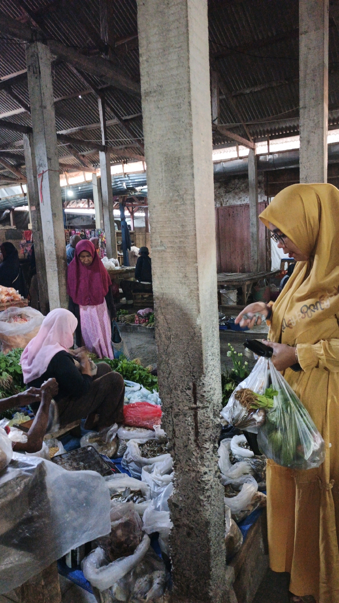 A group of people are gathered in what appears to be a market stall in Cibenda, Indonesia. In the foreground, a person wearing a yellow hijab and glasses holds a transparent plastic bag filled with vegetables and a bunch of green leafy produce. They are holding what looks like a phone in their left hand and appear to be interacting with someone off-camera. To the left, a person wearing a pink hijab is seated, facing away from the camera, with their hands in their lap. Another person, wearing a dark top and pink hijab, stands behind them, looking down. The market stall is characterized by its rustic architecture, with rough concrete pillars supporting a corrugated metal roof. Various goods, including produce and packaged items, are displayed on the ground, covered in plastic sheeting and bags. The overall scene suggests a bustling marketplace environment.