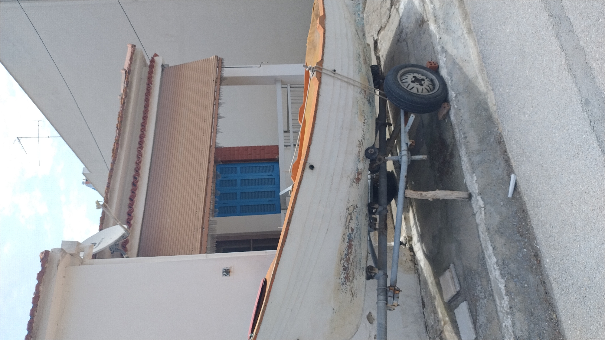 A white rowboat with an orange trim is parked on a trailer outside a building with a white facade and a brown retractable awning over blue shutters. The boat appears weathered, with peeling paint on its lower hull. The trailer has a single tire and is supported by a wooden post. In the background, a satellite dish is visible on the roof of an adjacent building, along with an antenna. The scene is located in Νέα Μηχανιώνα, Greece, on a paved surface next to a road. The sky is partly cloudy.