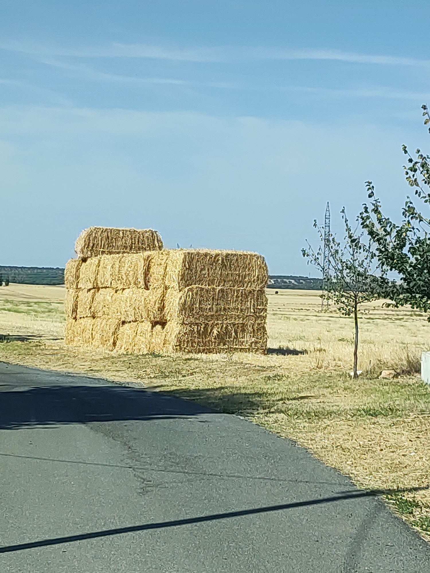 In Marugán, Spain, a rural scene unfolds under a clear blue sky with wispy white clouds. In the foreground, a paved road casts a long shadow across the parched earth. To the right of the road, a large stack of rectangular hay bales is neatly arranged, three layers high. Behind the hay, a field stretches out, its dry grass and stubble hinting at recent harvest. A solitary young tree stands near the hay bales, its branches sparsely leafed. In the mid-ground, a tall, lattice-like power pylon dominates the landscape. Further in the distance, a line of trees marks the horizon, with faint structures visible beyond them. The overall impression is one of a quiet, sun-drenched agricultural landscape.