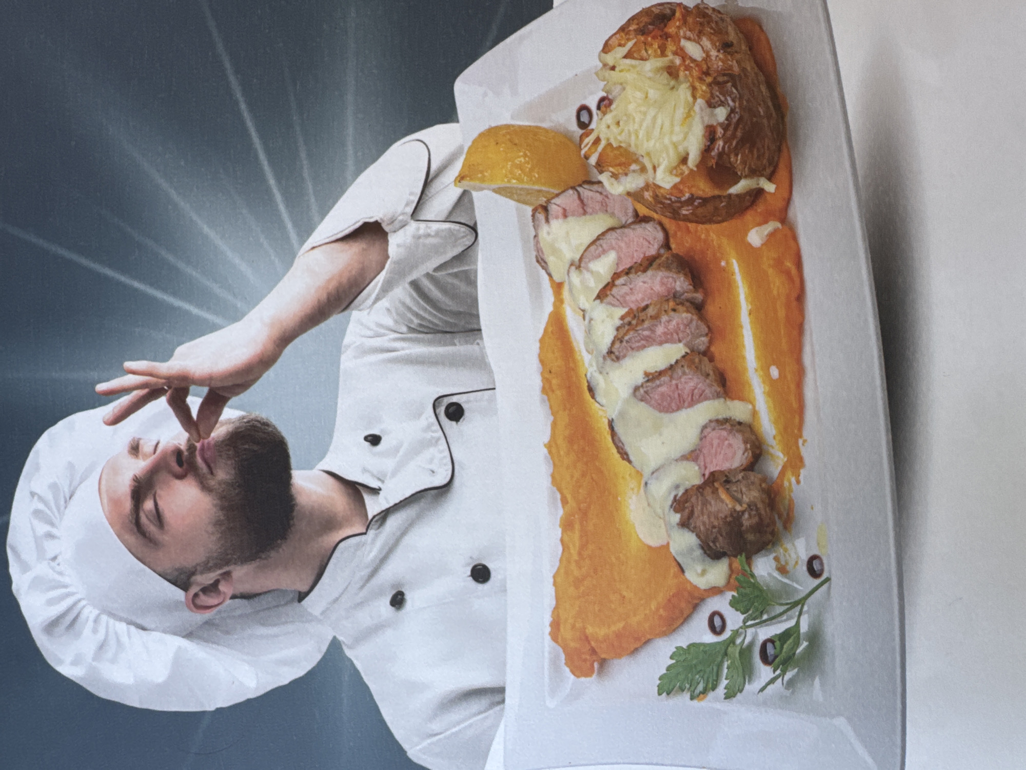 A professional-quality image, originally rotated 90 degrees counter-clockwise, features a male chef expressing culinary delight alongside a beautifully plated dish. The chef, a man with a beard wearing a white chef's coat with black buttons and trim, and a white chef's hat (toque), is positioned on the left side. His eyes are closed in apparent satisfaction as he performs the "chef's kiss" gesture, bringing his fingers to his mouth.
On the right, a long, rectangular white plate holds a gourmet meal. Sliced pieces of medium-rare meat, possibly pork tenderloin or beef, are arranged neatly and covered with a creamy white sauce. This protein rests upon a vibrant orange purée, likely sweet potato or carrot, artfully spread across the plate. Accompanying the meat is a large baked potato, split open and topped with a generous amount of melted cheese and some golden-brown elements, possibly nuts or fried onions. The dish is garnished with a bright yellow lemon wedge and a sprig of fresh green parsley, accented by a few small dark berries or peppercorns.
The background features a dramatic gradient, shifting from a dark blue-grey behind the chef to a lighter grey. Rays of light emanate from the upper left, behind the chef's head, creating a striking visual effect that highlights the subjects. The overall lighting is bright, emphasizing the textures and rich colors of the food. No text is visible in the image.
