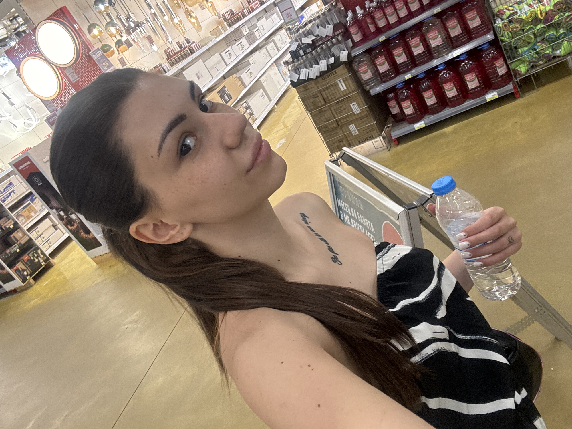 A female individual with long, dark brown hair styled in a ponytail is depicted in a slightly angled, eye-level indoor photograph taken in a large retail store in Sofia, Bulgaria. She has prominent eyebrows, discernible freckles on her cheeks, and her lips are slightly parted. She is wearing a black and white strapless top with an abstract stripe pattern. A script tattoo reading "She flies with her own wings" is visible on her left clavicle, and a small heart symbol is tattooed on her right hand near the thumb. Her fingernails are painted white with small, dark dot accents. She holds a clear plastic water bottle with a blue cap in her right hand, positioned in the lower center of the frame.
The background displays a commercial retail environment with a tan-yellow floor. To the upper left, shelves are stocked with various boxed items, and a red sign partially displays the text "GAUTENG" and "BULGARI" or "BULGARIA". A white sign below it shows "MEZELER / BAL". To the upper right, multiple lighting fixtures are mounted and displayed, including two large, illuminated circular ceiling lights. In the mid-ground to the right, shelves are stacked with numerous large red plastic bottles, likely beverages. Below these bottles, a wire basket contains colorful, bundled merchandise. Directly behind the subject, cardboard boxes labeled "LIGHTEX" are stacked. The scene suggests a department within a large home improvement store or hypermarket, featuring lighting products, beverages, and other consumer goods.