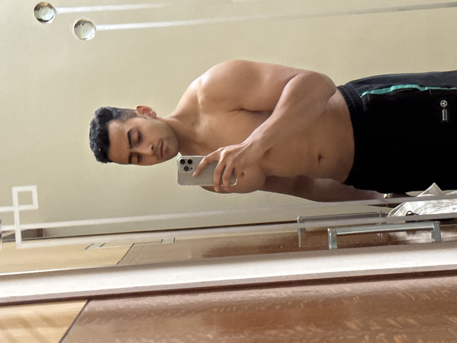 An adult male is depicted taking a mirror selfie indoors in Kolkata, India. He is shirtless, from the waist up, and wears black track pants featuring a teal stripe on the outer seam of the left leg and a small logo near the left hip. The individual holds a silver smartphone with a triple camera system in his right hand, pointed towards the mirror. He has dark, short hair with faded sides, dark eyebrows, and a subtle mustache. The background in the mirror reflection consists of a light-colored wall adorned with two vertically aligned circular, clear decorative objects and a white angular geometric pattern etched onto the mirror surface. To the left of the reflected subject, a wooden door or wardrobe with a metallic handle is partially visible, with a white garment hanging behind it.
