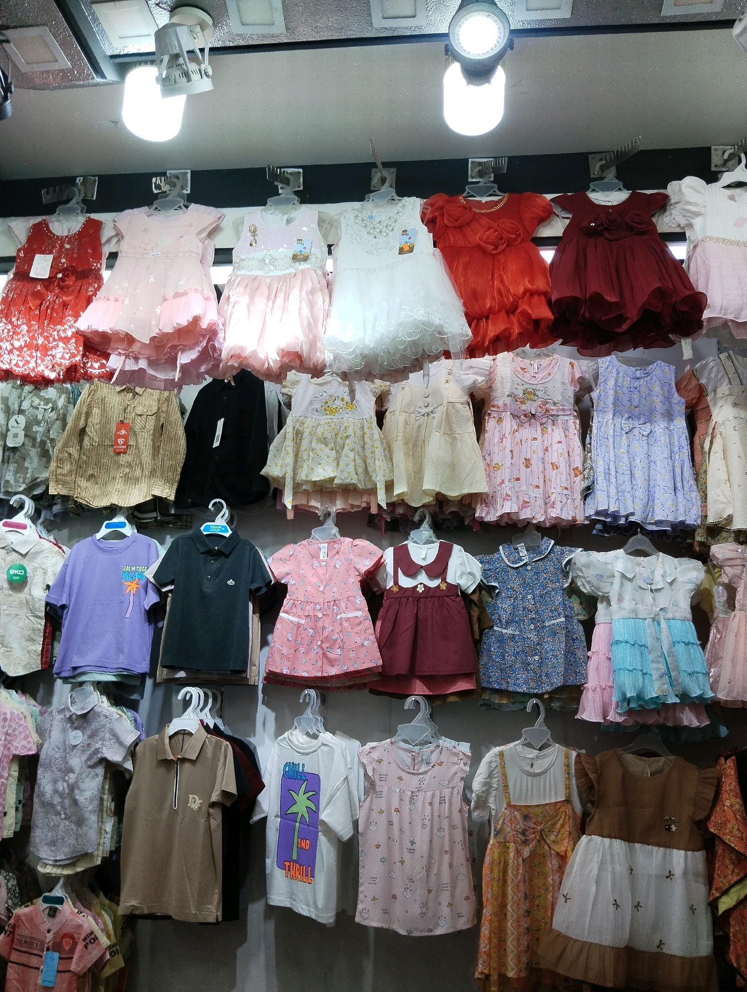 A stall displaying a variety of children's clothing is shown in an indoor market in Noakhali, Bangladesh. The clothing, primarily dresses and shirts, is hung on hangers from a rack above. The items are arranged in multiple rows, with some clothing items folded and stacked on shelves below. The stall is illuminated by artificial lighting. The image focuses on the merchandise, with no people visible. The background consists of a gray wall and what appears to be a ceiling with lighting fixtures.