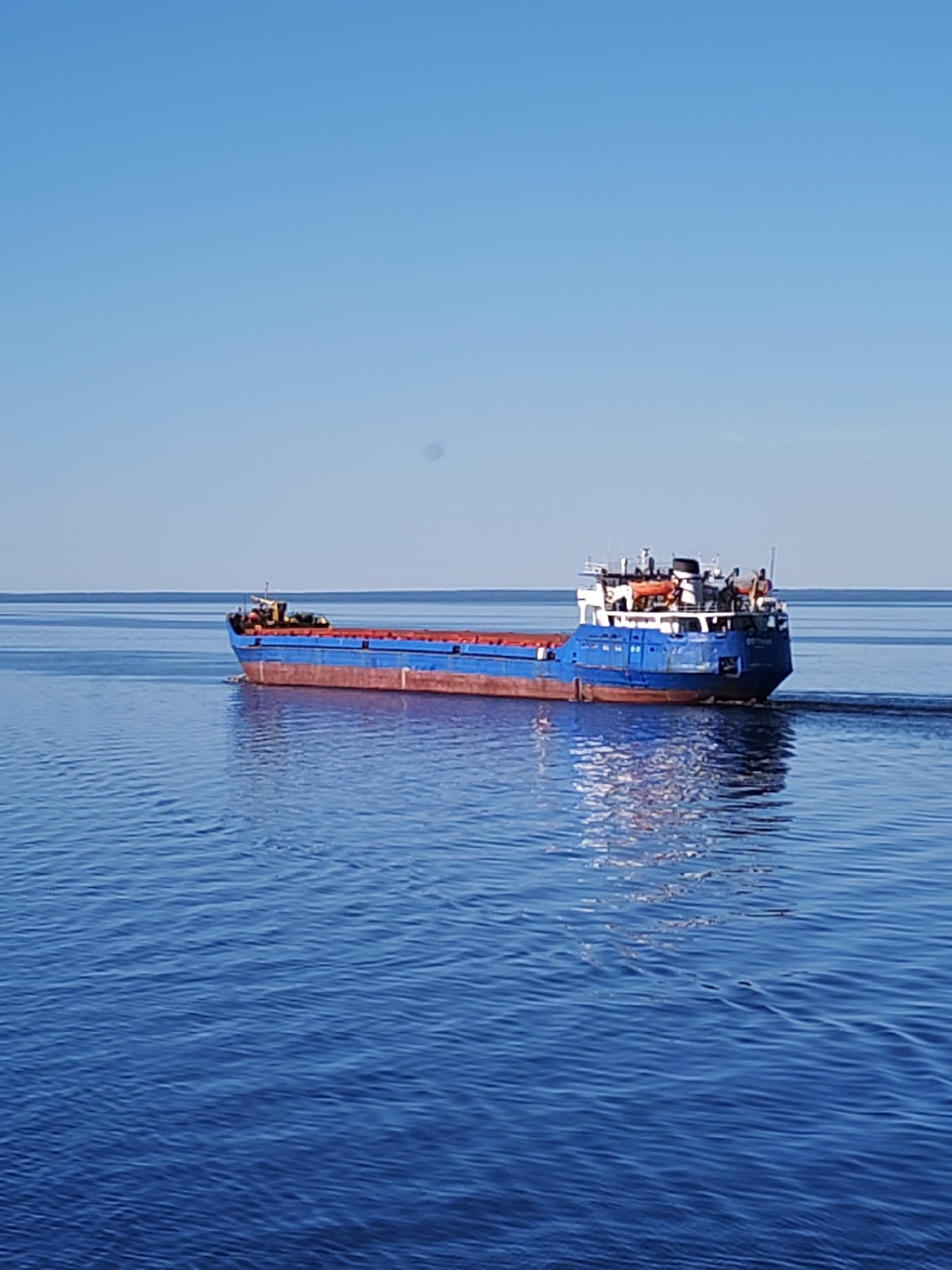 An empty blue and reddish-brown cargo vessel named "ОСТИН" (OSTIN) traverses calm, deep blue water under a clear, light blue sky in the vicinity of Ермаково, Russia. The ship is moving from left to right, indicated by a visible wake trailing from its stern. The vessel features a reddish-brown hull below the waterline and a blue hull above, with a multi-decked white superstructure capped in black located at the stern. Several indistinct figures, likely crew members, are visible on the superstructure. The water reflects the blue sky, with a flat horizon line visible in the distance. The overall scene depicts bright daylight conditions with minimal atmospheric haze.