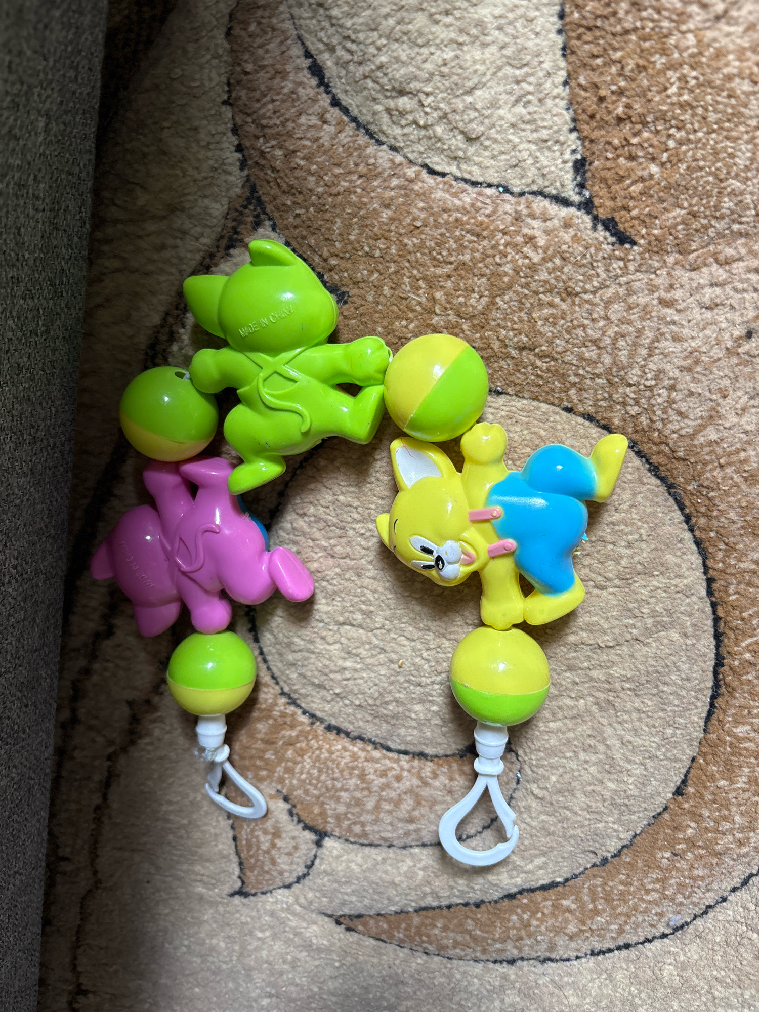 A high-angle, close-up shot shows three plastic baby toys scattered on a brown and tan carpet. The toys are a green alien-like creature, a pink elephant, and a yellow and blue cat-like character. Each toy is attached to a round rattle that is half green and half yellow, with a white plastic clip for attachment. The green toy is on the top left, the pink elephant in the middle left, and the yellow and blue cat on the right. The toys appear to be part of a baby crib mobile. The carpet has a swirling pattern of dark brown and tan fibers. The scene is indoors, likely a child's room, and there are no people or animals visible. The image does not provide any information that would allow for location identification in Sinegorskiy, Russia.