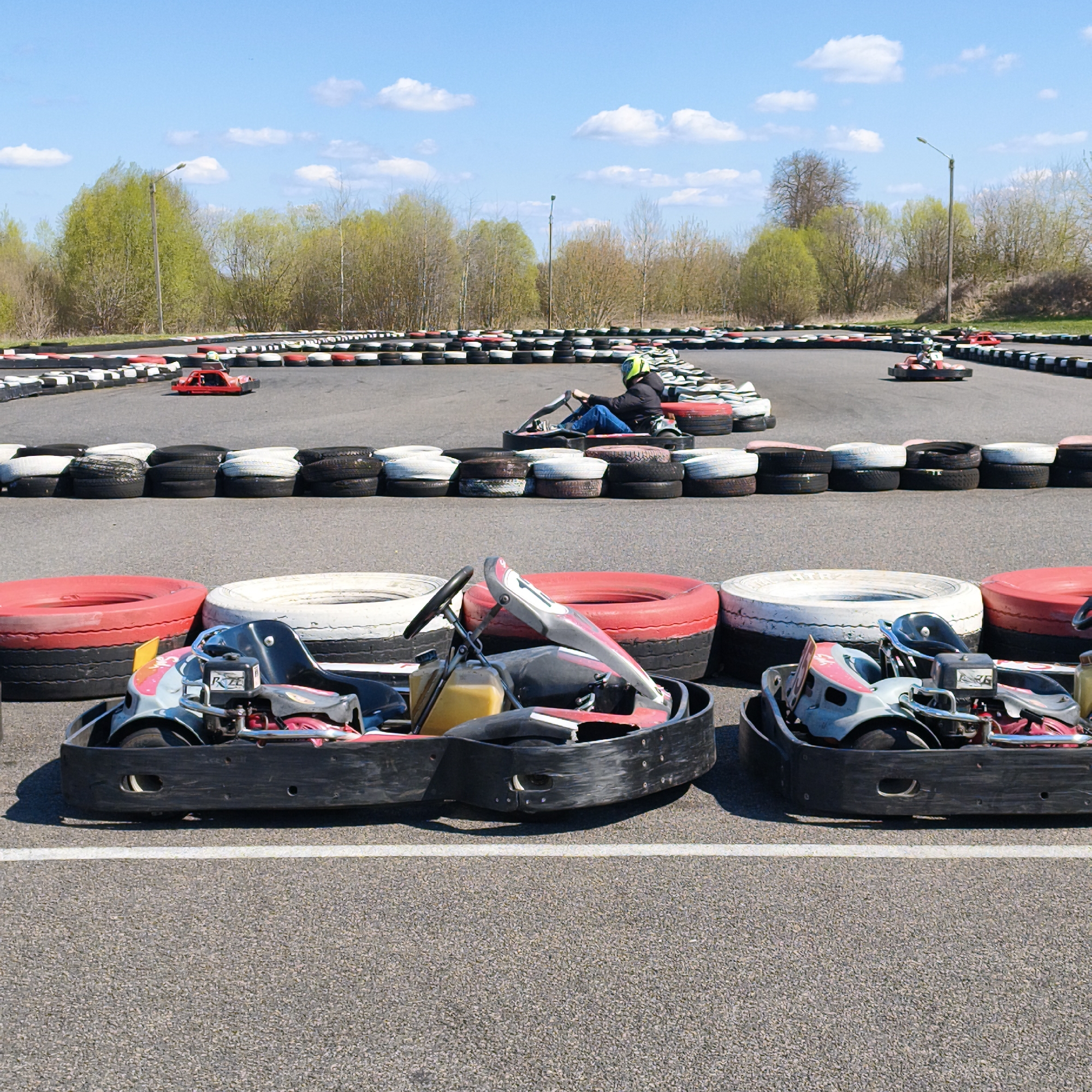 The image depicts a go-kart track located in Hrodna, Belarus. Several go-karts are visible. Two are parked in the foreground on the paved track surface, separated by a white painted line. In the background, one go-kart is in motion on the track with a person seated. Additional karts are stationary along the track. The track's perimeter is delineated by rows of stacked tires, painted black and white, and red and white. In the background are deciduous trees under a blue sky with scattered clouds. Two street lights can be seen. The track surface is asphalt.