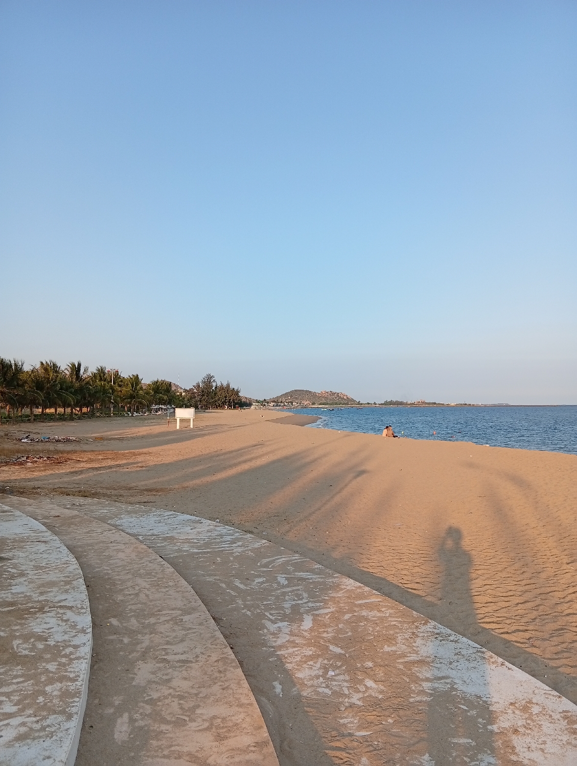 This media file captures a serene coastal landscape in Khánh Hải, Vietnam, likely during late afternoon or early morning due to the warm light and long shadows. The sky is a clear, cloudless blue, indicating fine weather.
In the foreground, a curved concrete structure, resembling a walkway or stepped seating, frames the left and bottom edges of the image. The shadow of a person, presumably the photographer, is cast prominently across the sand in the lower right.
The central part of the image features a wide expanse of golden sandy beach, stretching towards the distant horizon where it meets calm, blue-green ocean waters. Several distinct, elongated shadows, likely cast by unseen palm trees or other structures behind the camera's view, stretch diagonally across the sand. Two individuals are visible in the mid-distance, seated or reclining on the sand close to the water's edge. Further along the shoreline, a line of palm trees and dense foliage borders the beach, with a small, white rectangular sign or shelter visible among the greenery.
The water itself appears tranquil, with a few small boats discernible in the distance. Low hills or mountains rise faintly on the far horizon beyond the coastline, completing the scenic view. No visible text is apparent in the image. The scene evokes a sense of peace and relaxation by the sea.