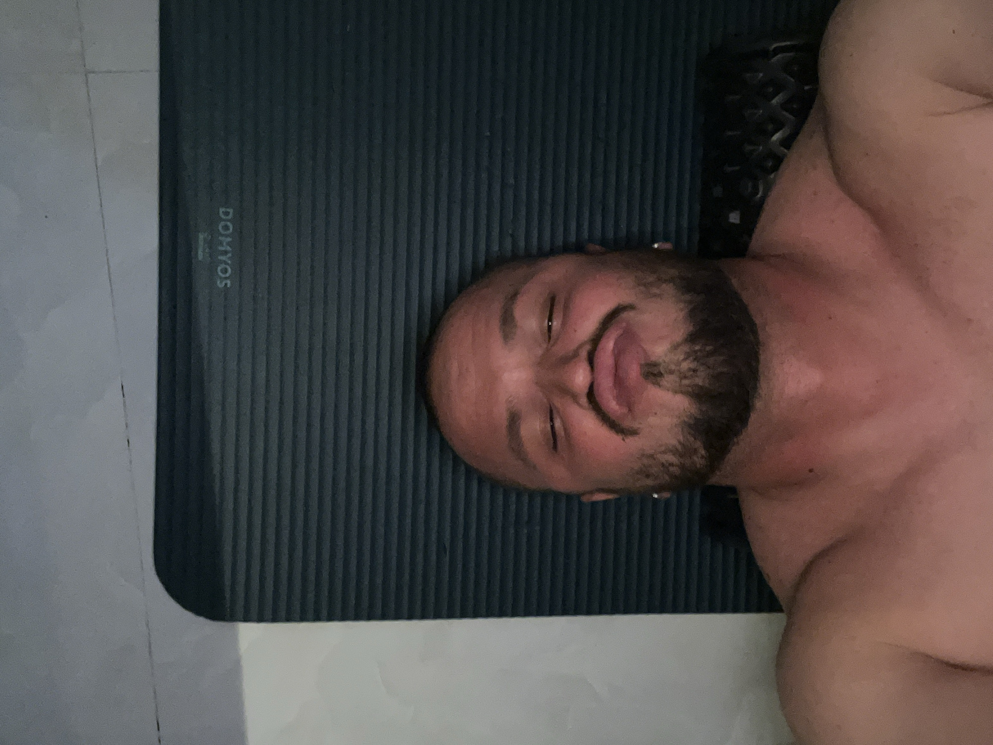 A shirtless, bald man with a beard and a small hoop earring in his left ear puckers his lips and closes his eyes as he holds the camera up for a selfie. He is lying down on a dark blue yoga mat with horizontal ridges. The mat is placed on a light-colored, tiled floor. The background is blurry, but appears to be a wall with some texture. The man's shoulders and upper chest are visible. The lighting is dim, with a reddish hue that may indicate a flash. The word "DOMYOS" is visible in white lettering on the yoga mat.