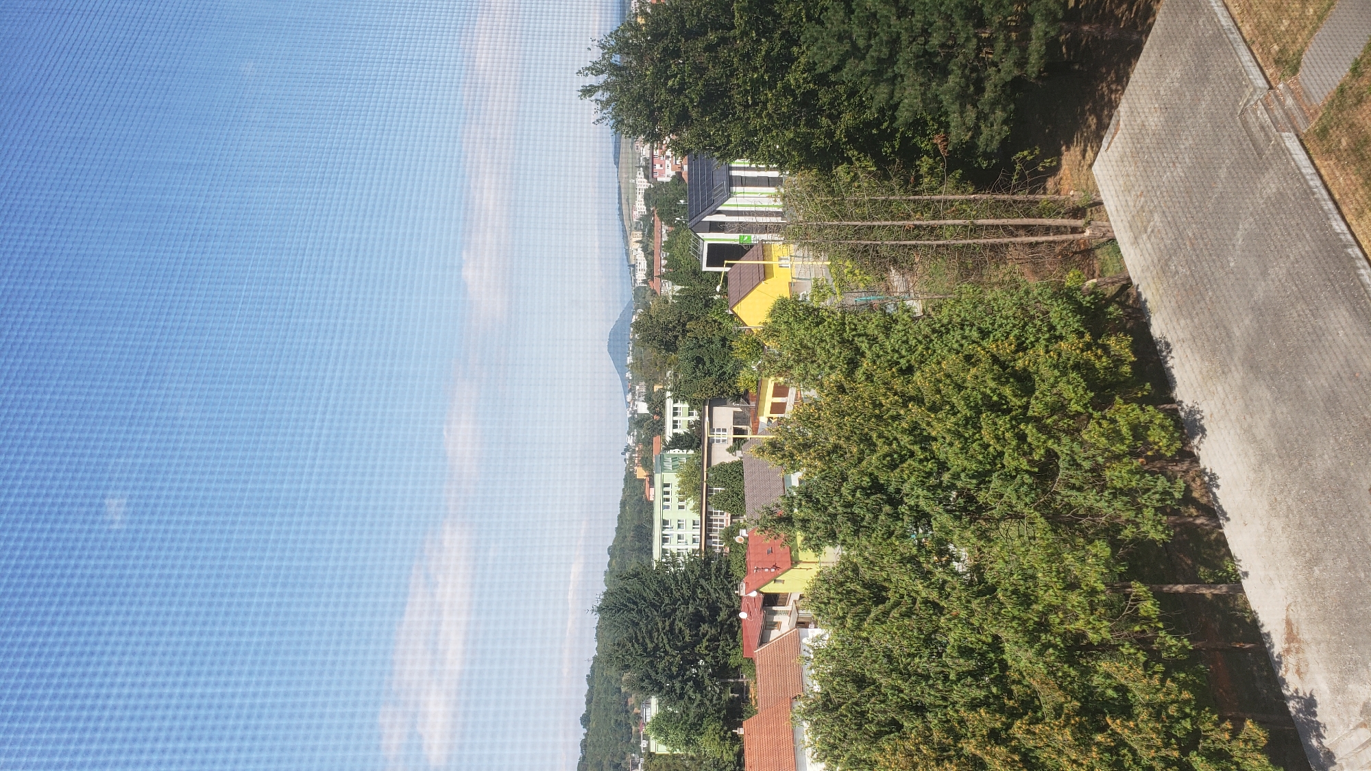 This is a high-angle, full shot of a residential area in Prešov, Slovakia, viewed through a window with a screen pattern. The sky is clear blue with a few wispy clouds. In the foreground, a paved area with concrete slabs is visible. Behind this, a row of tall evergreen trees forms a dense barrier, with some of their branches showing signs of autumnal color change.
Beyond the trees, a collection of houses and buildings are nestled together. Several houses have red-tiled roofs, while others are painted in yellow and green. One distinctive building has a modern design with green and black striped panels and solar panels on its roof. In the middle ground, a larger, light green building with multiple windows stands out.
Further in the background, a range of hills and mountains can be seen, with a prominent, conical-shaped mountain peak visible on the left side. The urban landscape extends towards the horizon, with a mix of residential buildings and some taller structures. The overall impression is of a suburban or outer-city residential neighborhood on a clear day. No people are visible in the image.